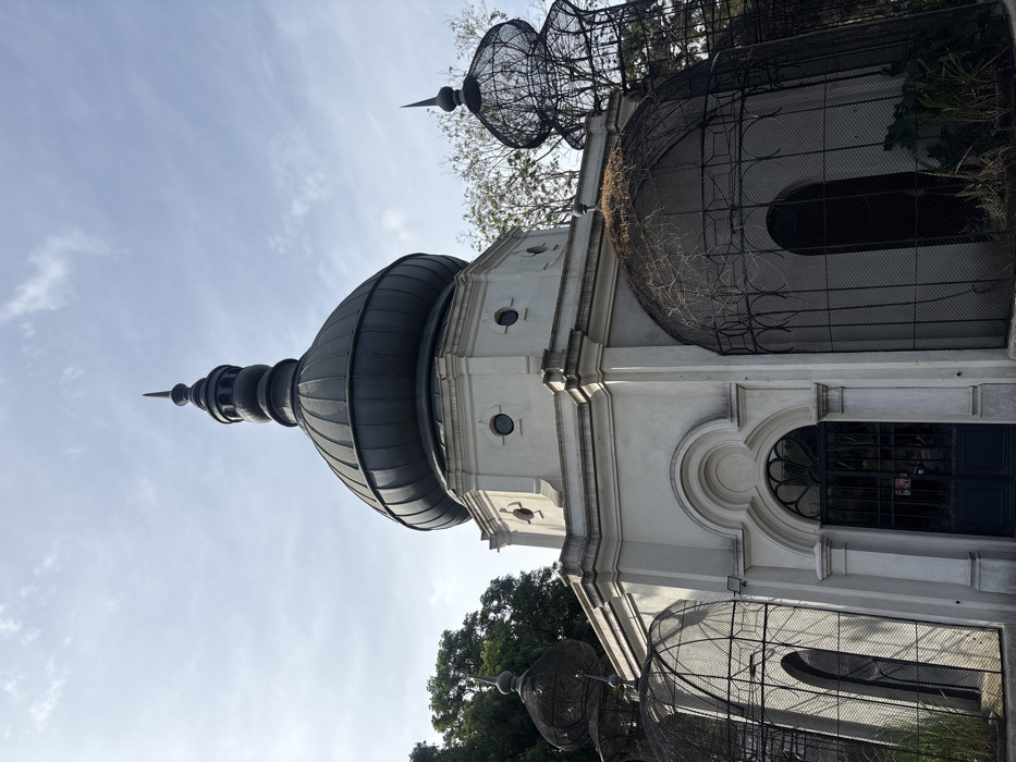 The image displays the Pabellón de los Cisnes (Swan Pavilion) located within the Jardín Botánico Carlos Thays in the Palermo neighborhood of Buenos Aires, Argentina. The central structure is a multi-tiered, light-colored octagonal building topped with a large, ribbed, dark grey onion-shaped dome and a pointed finial. The building's lower section features an arched main entrance with dark double doors, on which a small red sign is affixed, and a security camera mounted above. The mid-section, or drum, presents small circular windows (oculi) and decorative architectural elements. Flanking the central pavilion are several large, dark metal, dome-shaped wire mesh structures or aviaries, some partially covered by dry vines, also featuring pointed finials similar to the main dome. Lush green trees are visible in the background and sparse ground vegetation around the base of the structures. The sky is partially cloudy blue. No individuals are present in the scene.
