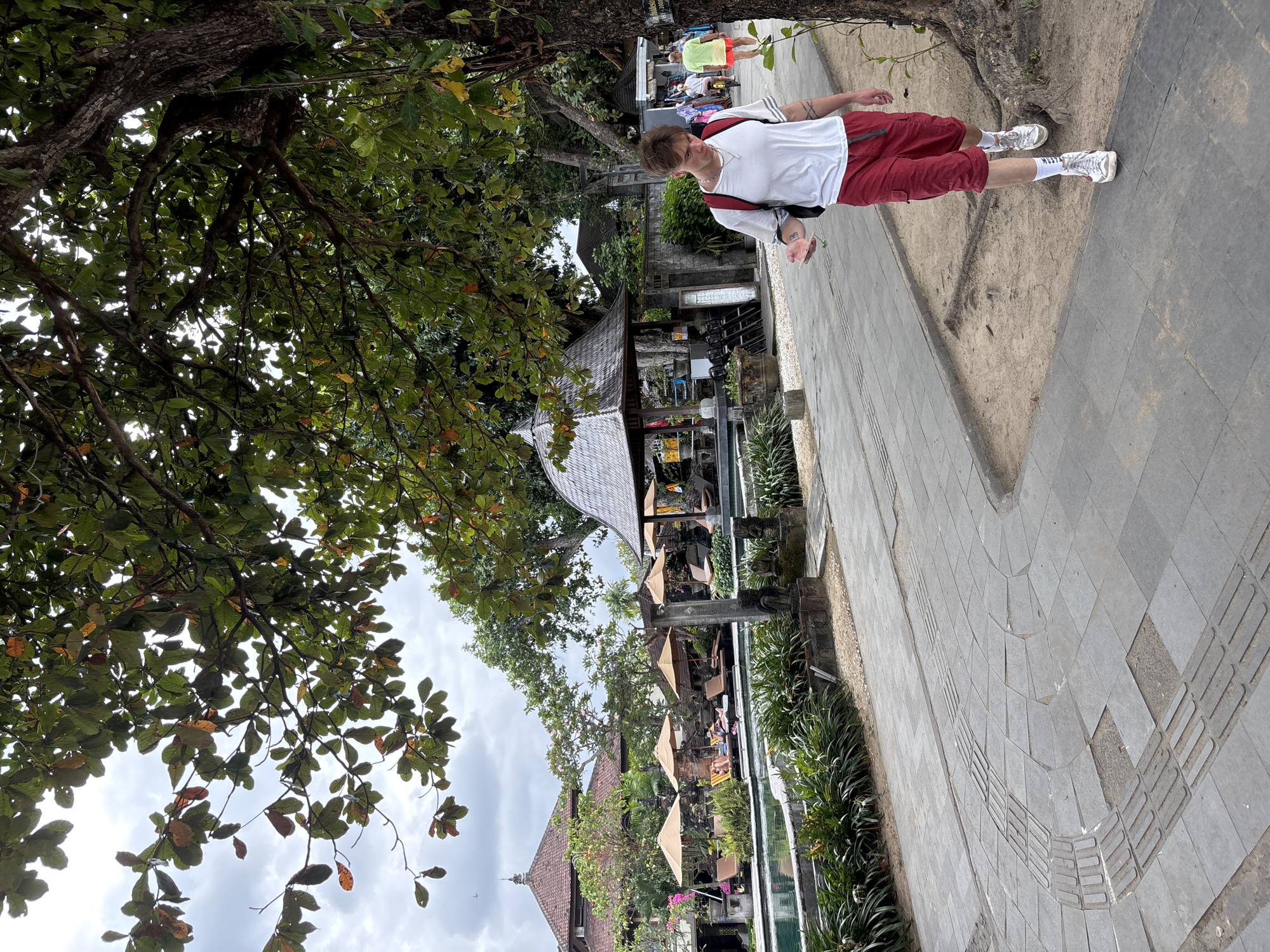 A young man with short brown hair, wearing a white t-shirt, red cargo shorts, and white sneakers, walks along a paved path. He has a red backpack on his shoulders and appears to be holding a small object in his left hand. To his left, there is a swimming pool with lounge chairs and umbrellas. In the background, there are buildings with traditional Indonesian architecture, including a red-tiled roof and a pavilion with a thatched roof. Lush greenery and trees surround the area. The sky is overcast. This scene is likely located in a resort or hotel in Denpasar, Indonesia, given the architectural style and tropical vegetation.