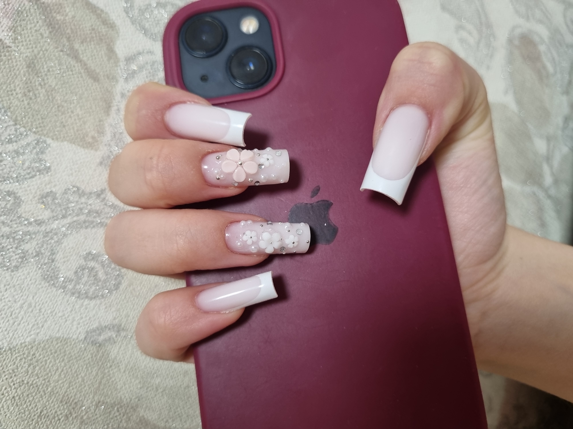 A close-up shot shows a hand holding a burgundy-colored phone. The fingernails are long and square-shaped with a white French manicure. One nail is decorated with a row of small white beads, and another has a pale pink flower design with surrounding beads. The phone has an Apple logo visible. The background appears to be a patterned, possibly textured, fabric or surface. The location is not identifiable as Chaikovsky, Russia, based on the visual information provided.