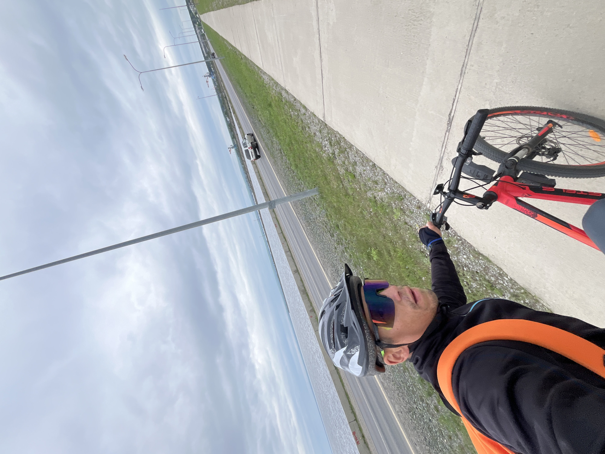 The image shows a first-person perspective of a man on a red bicycle, wearing a black jacket, helmet, and reflective sunglasses. He is on a paved sidewalk next to a road with cars. The road runs alongside a body of water and a cloudy sky. In the background, industrial structures and streetlights are visible. This scene appears to be located in Chaikovsky, Russia, likely along the Kama River or one of its tributaries, given the presence of a large body of water and industrial elements often found near such waterways in Russian cities. The man is actively cycling, holding the handlebars with his right hand.