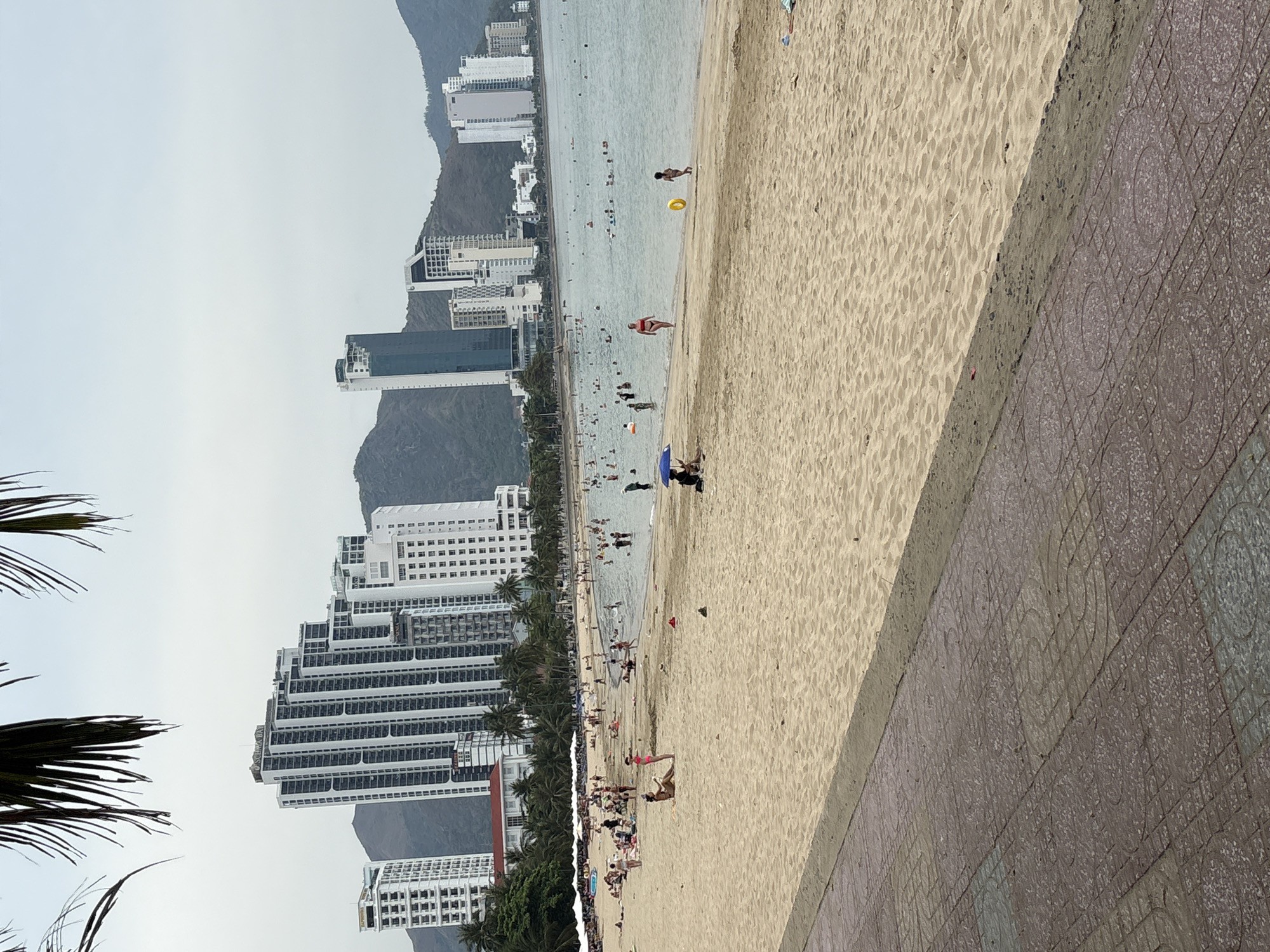 This image shows a beach in Bac Nha Trang Ward, Vietnam. The shoreline curves gently, revealing a sandy beach occupied by numerous individuals. Some people are swimming in the water while others are relaxing on the sand. There are several high-rise buildings on the left, some of which are located on a steep hillside. In the foreground there is a sidewalk bordering the beach.