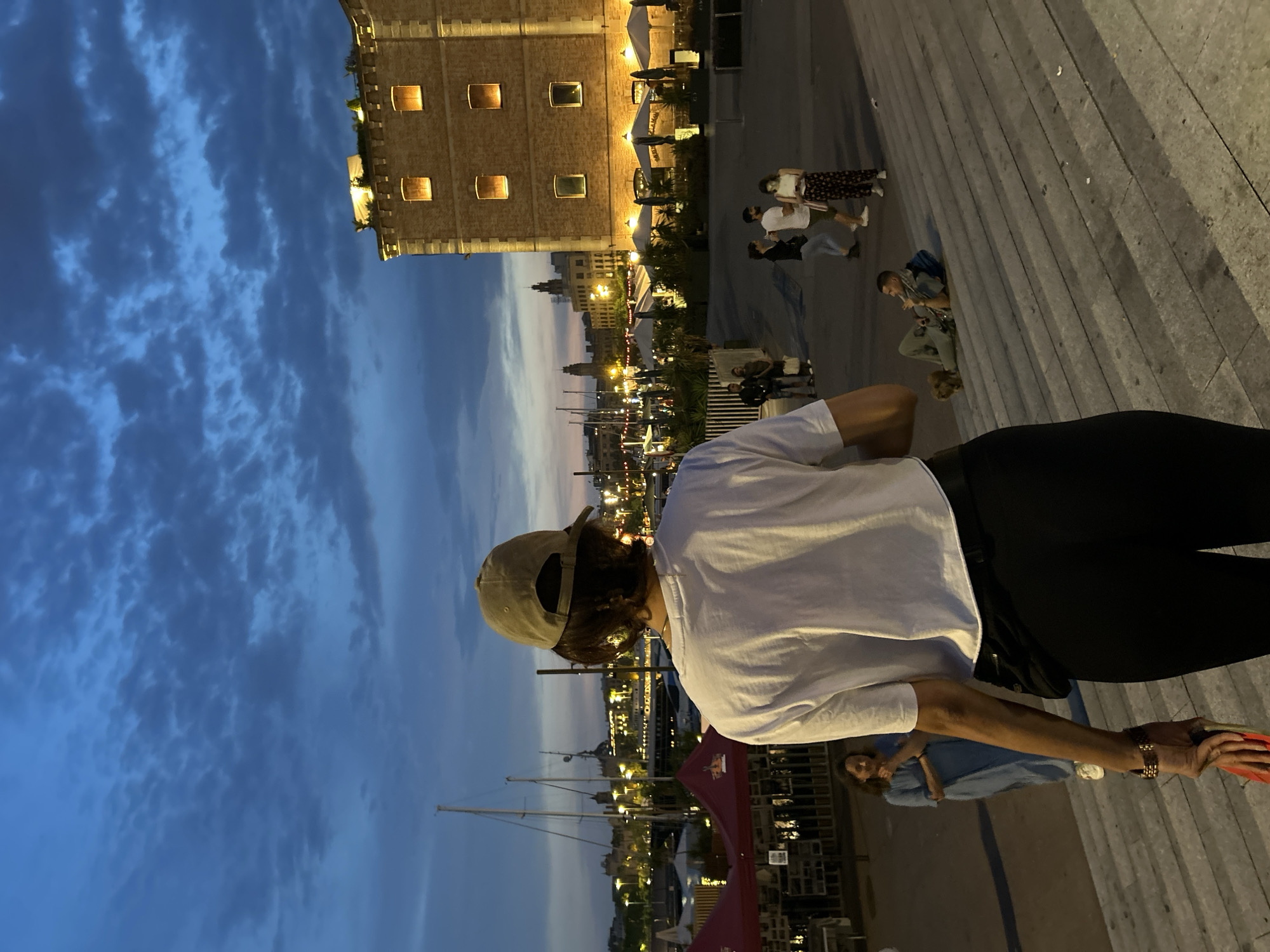 A photo taken at dusk depicts a person wearing a white T-shirt, black pants, and a baseball cap. The person is standing on a stone staircase overlooking Barcelona harbor. The Moll de la Fusta, a waterfront promenade with bars and restaurants, is visible in the background. Sailboats are docked in the harbor. The sky is a mix of blue and gray clouds. In the far background, the illuminated Port Authority building rises in the distance. A few people are seated or strolling along the promenade, and others are on the staircase. The scene is Barcelona, Spain.