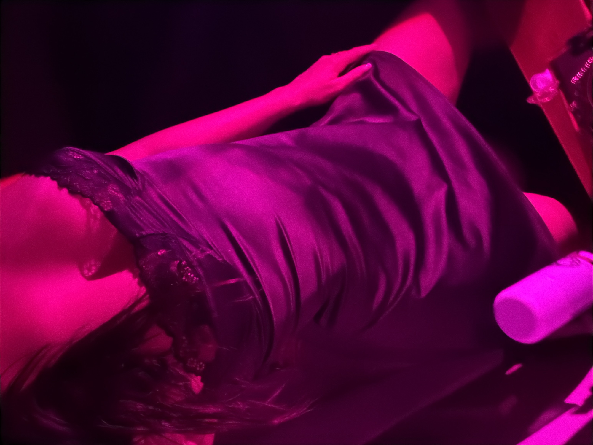 A high-angle close-up shot depicts a person reclining, possibly in a bedroom, under a pink-purple light that dominates the scene. The subject, seen from the shoulder up, is wearing a dark, satiny garment trimmed with lace, with a hand resting gently on the fabric. The person's dark hair is spread across the surface they're reclining on, and part of their face is visible, although obscured by shadow and the overall color cast. To the right, a cylindrical object, possibly a water bottle, and a table with various items are partially visible. The environment appears to be dimly lit, contributing to the intimate, almost dreamlike, quality of the image. The location and city cannot be determined from the image.
