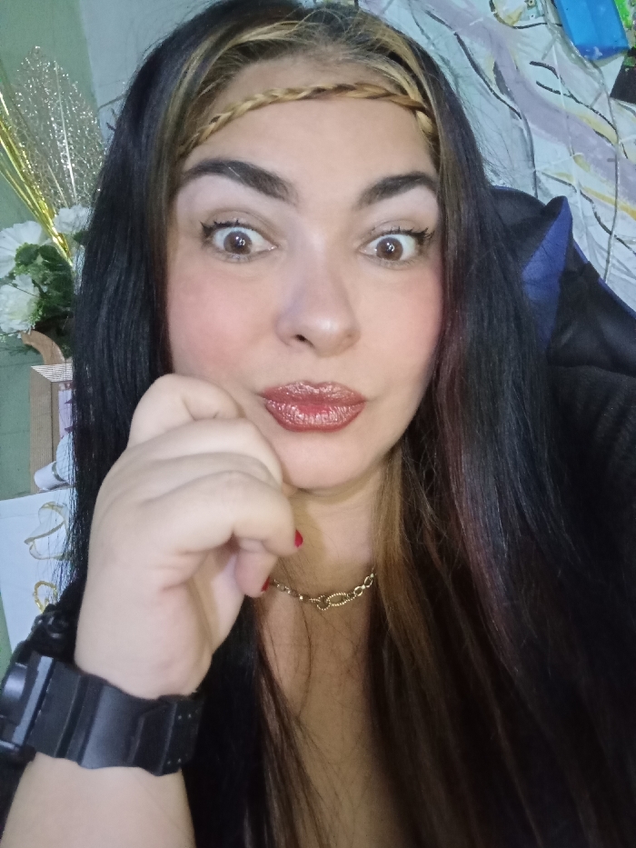 A woman with dark hair and a braided headband looks directly at the camera, her eyes wide. She has fair skin, red lipstick, and her hand is near her cheek in a thoughtful pose. She wears a gold necklace and a large black wristwatch. In the background, there are decorative white flowers and green leaves, along with a patterned sheet or curtain.