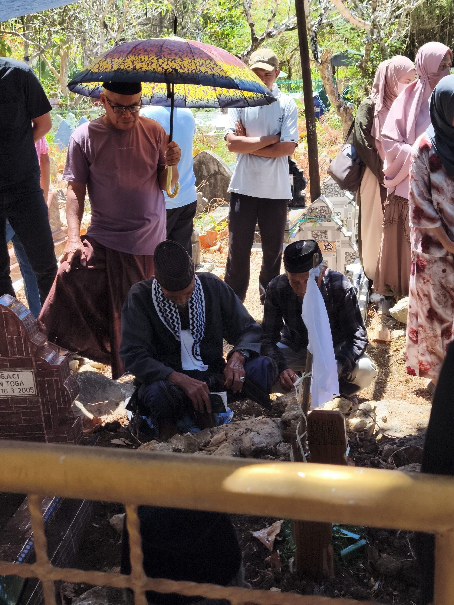 The image depicts a funeral procession in the city of Tonasa, Indonesia. In the foreground, two men are kneeling near a grave. One man, wearing a black cap, black shirt, and a checkered scarf, is looking down at something in his hands. The other man, wearing a purple shirt and a dark skirt, is holding an umbrella over his head.
In the background, several women are standing, all wearing headscarves in various colors. Some are looking towards the grave, while others are looking away. A young man in a white polo shirt and a brown cap stands with his arms crossed, observing the proceedings. The environment appears to be an outdoor cemetery, with several graves visible in the background. The ground is covered with dry leaves and rocks. The overall atmosphere seems solemn and respectful.