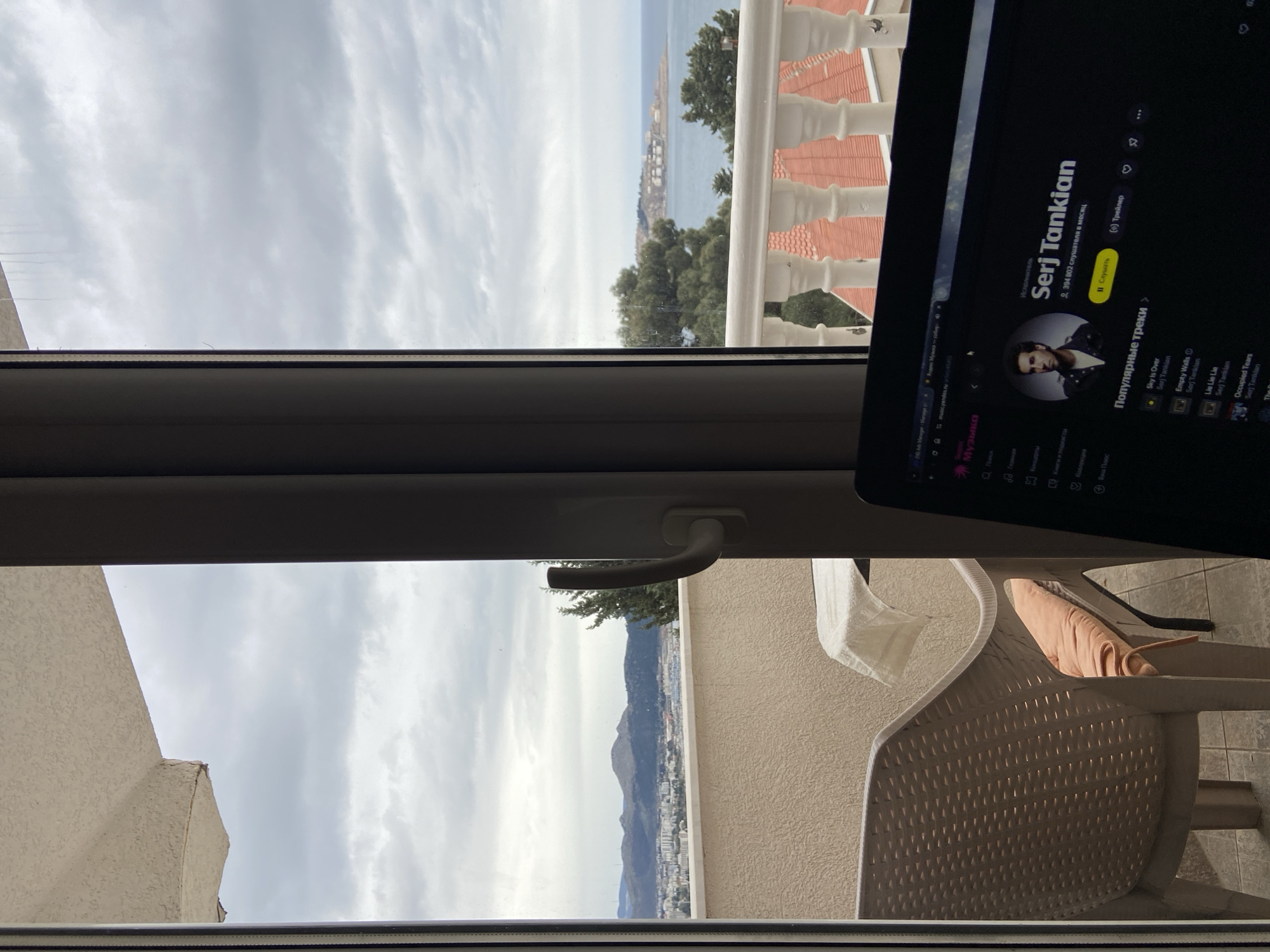 The image depicts an interior view through a window. Outside, a cloudy sky hangs over a distant coastal cityscape, potentially Šušanj, Montenegro, nestled along a bay or sea. In the foreground, a laptop screen displays the artist Serj Tankian on a music streaming service. A white plastic chair sits partially visible to the right, with a light-colored fabric draped over it. A window handle is also visible.