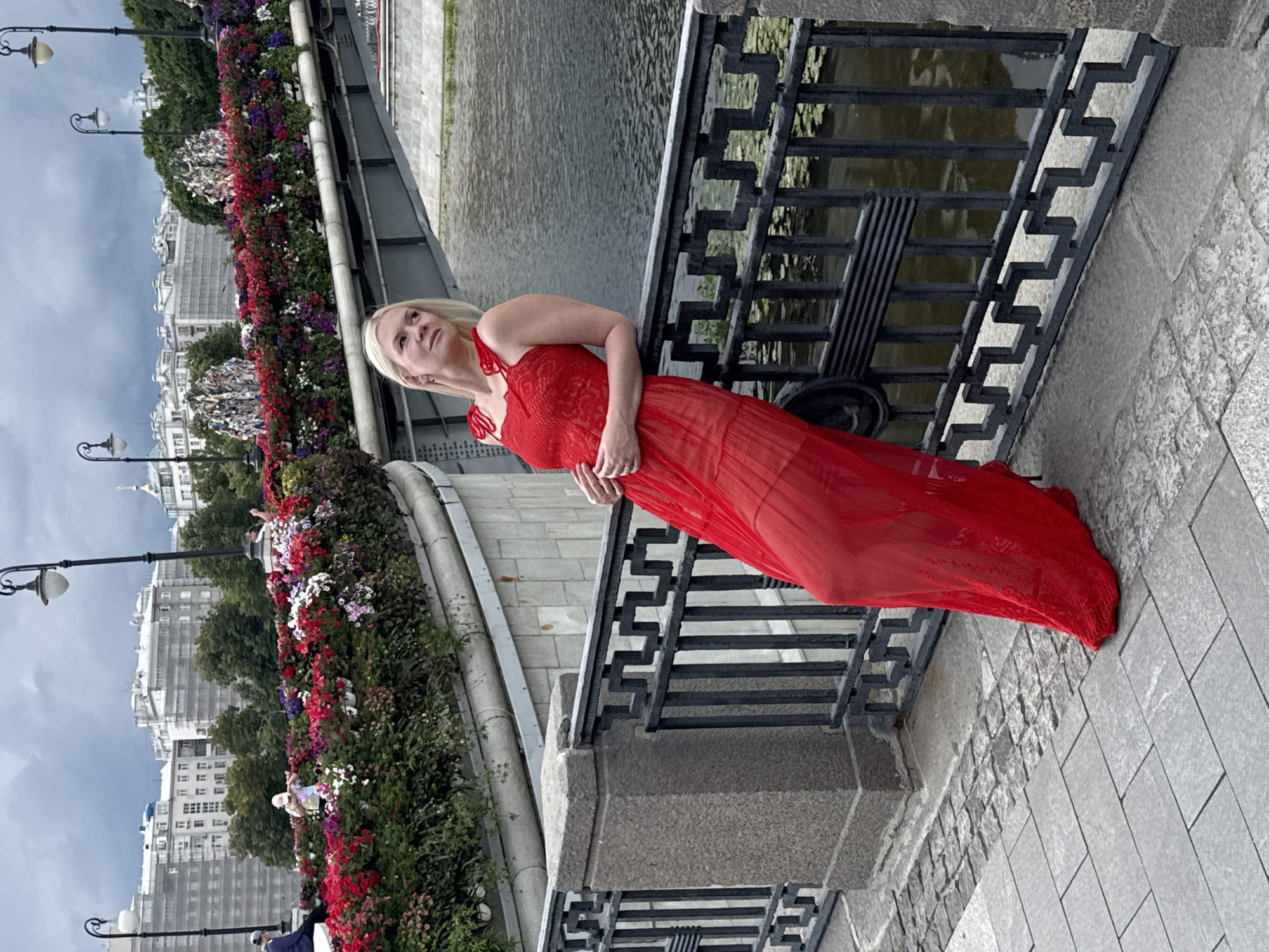 A woman with blonde hair, wearing a bright red, sheer, floor-length dress with spaghetti straps and a textured bodice, leans against a black metal railing on a stone embankment. Her left hand rests on her hip, and her right arm is bent at the elbow. She looks upwards and to her left, with her head tilted back slightly.
The railing she leans on is ornate, with a geometric pattern. Behind her, a wide, grey river flows, with green aquatic plants visible on its surface. A bridge spans the river, its underside and supports visible. The bridge is adorned with a thick band of colorful flowers – reds, pinks, purples, and whites – in tiered planters. Beyond the bridge, city buildings with white facades and scaffolding are visible under a cloudy, grey sky. Lampposts with traditional lantern-style lights are scattered along the bridge and embankment. The ground in the foreground is paved with cobblestones and large stone slabs. The overall impression is of an urban scene with a prominent natural element (the river and flowers) and a focus on the woman. The presence of scaffolding suggests ongoing construction or renovation in the city. This scene is identifiable as the Moskva River embankment near the Patriarchal Bridge in Moscow, Russia, given the distinctive architecture and the common practice of decorating bridges with flowers.