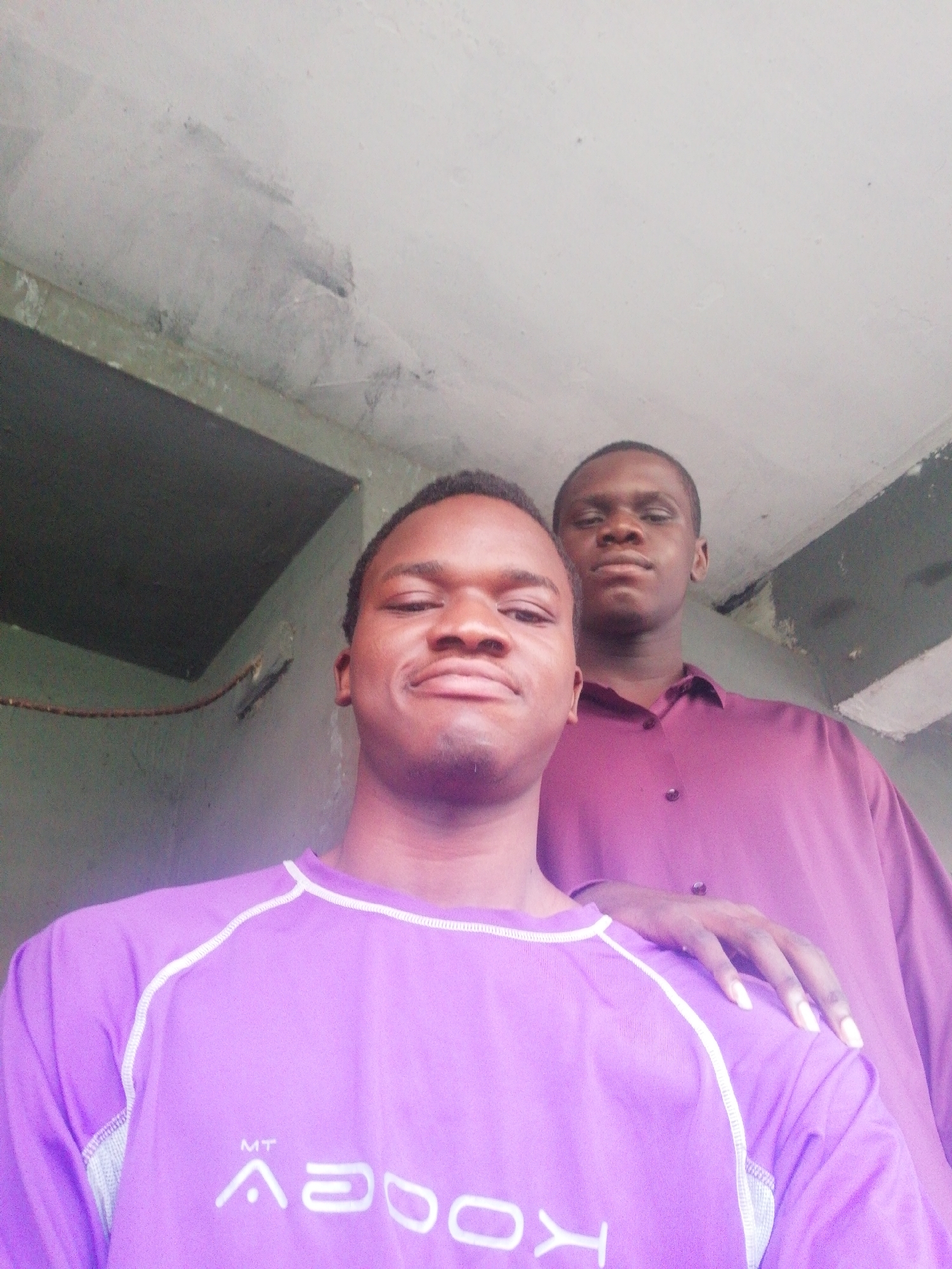 Two young men are posing for a photo. The man in the foreground wears a light purple shirt with white trim and white lettering that reads "MT ADOOV." The second man stands behind him, wearing a maroon button-down shirt. The second man has his hand on the first man's shoulder. The location appears to be outdoors, possibly near a building with a gray exterior. The setting is likely in Enugu, Nigeria.
