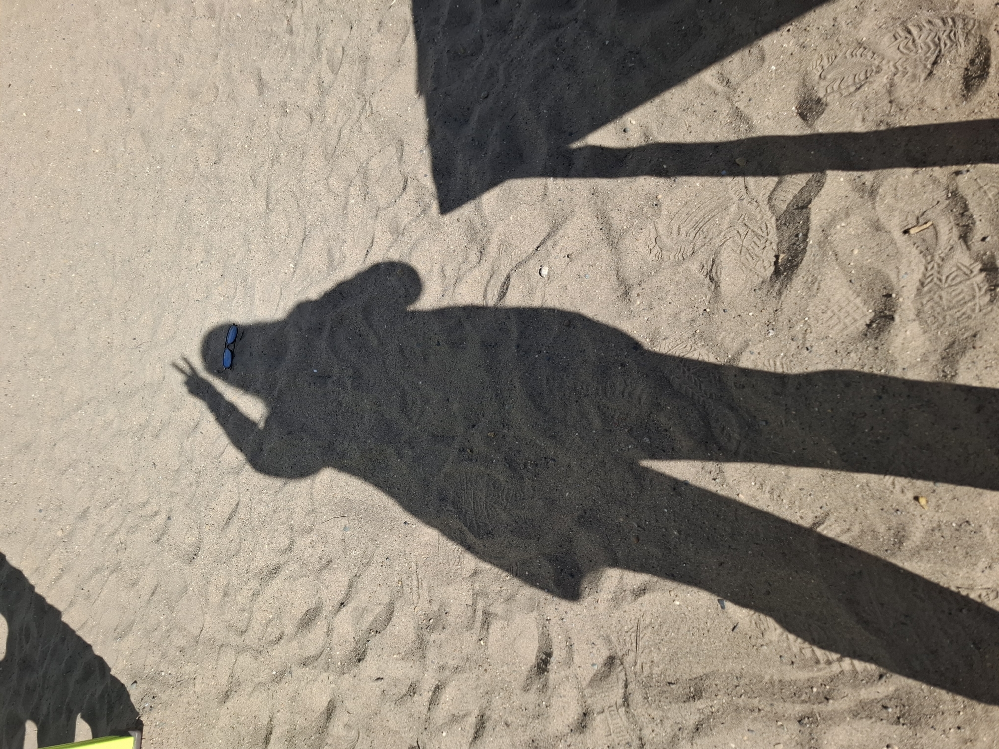 The image shows an overhead view of a sandy area, likely a beach or playground, with shadows cast upon it. A shadow of a person, likely the photographer, is prominent, with the person's shadow making a peace sign with their fingers. A pair of sunglasses rests near the shadow's head. Other shadows, possibly from nearby structures, are also visible. The sand shows scattered footprints. The specific location is unknown, although it is said to be in Chaikovsky, Russia.