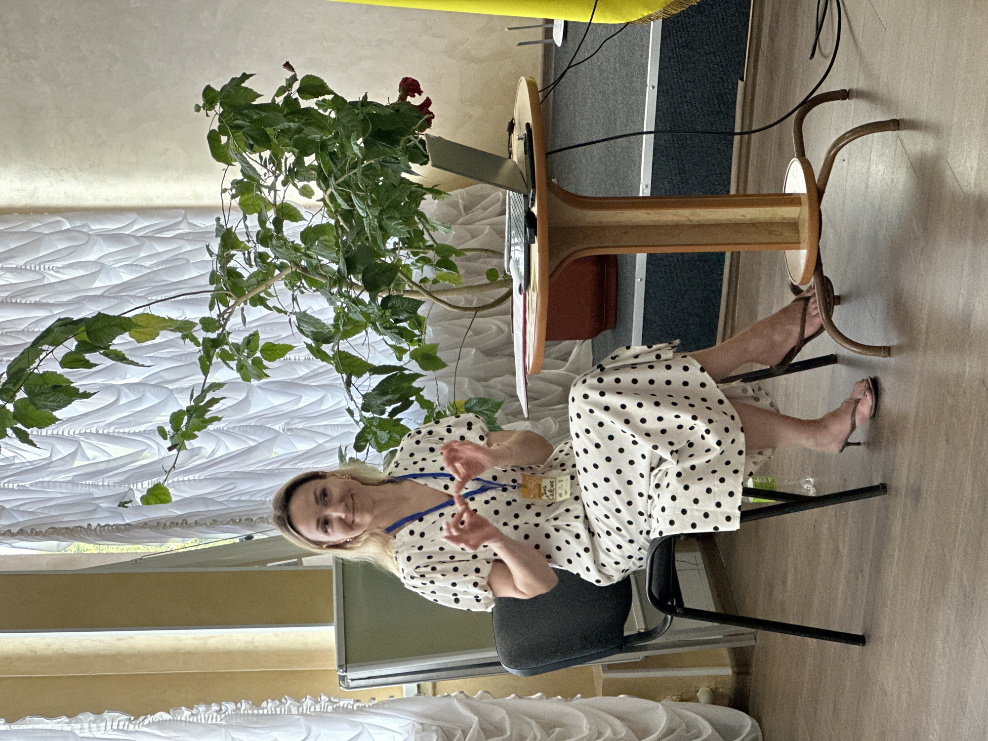 A woman with blonde hair is sitting in a black chair in what appears to be a conference room or lecture hall in Myronivka, Ukraine. She is wearing a white dress with black polka dots and has a lanyard around her neck with a badge. The woman is gesturing with her hands as if speaking. In front of her is a round wooden table with a laptop, a phone, and some papers. A large potted plant with green leaves is positioned behind the table. The room has white curtains in the background and a yellow flag on the right side. The floor is made of dark grey carpet and wooden planks.