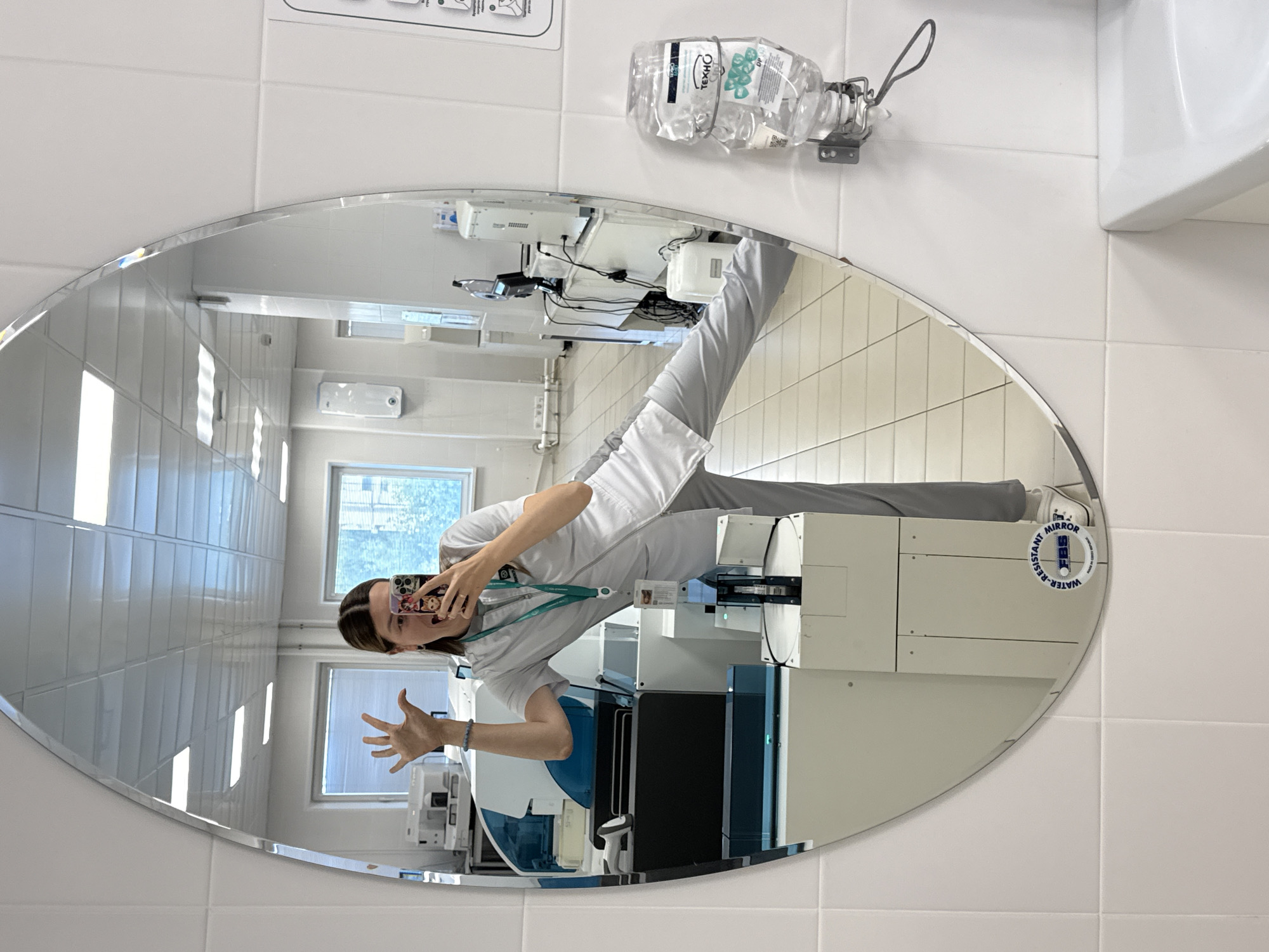 A woman is taking a mirror selfie in what appears to be a laboratory or medical facility. She is wearing a light grey uniform and has her left hand raised with fingers spread. She is holding a smartphone in her right hand, which is partially obscuring her face as she takes the photo. The reflection shows a laboratory setting with medical equipment, including what looks like a blood analyzer, and tiled walls and flooring. A dispenser of hand sanitizer is mounted on the wall to the right of the mirror.
