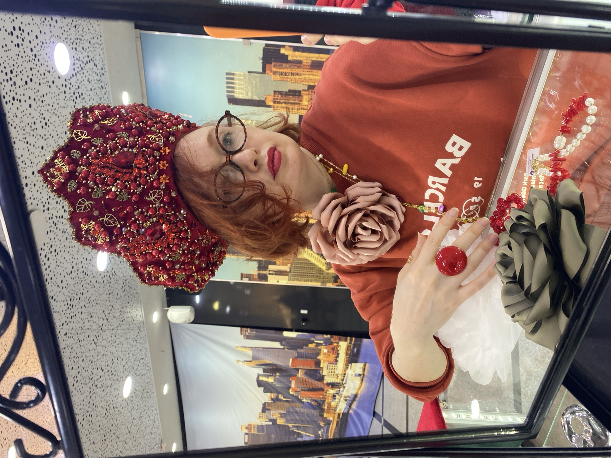 This image captures a reflection, likely in a mirror or glass display case, of a woman in an indoor setting in Yekaterinburg, Russia. The woman has reddish-orange hair and fair skin, with her lips painted a dark red. She wears round, tortoise-shell rimmed glasses and an orange-red long-sleeved sweatshirt. Adorning her head is an elaborate, ornate red headpiece, richly decorated with numerous red beads, jewels, and gold-colored leaf patterns. Around her neck, she wears a necklace that features a large, light pink fabric rose and a string of yellow and white beads. On her left ring finger, a prominent, large red ring is visible.
Her hand is positioned near various items within the display, which include a necklace or bracelet composed of red coral-like beads and white pearls, another red beaded accessory, and a folded item of dark olive-green fabric, possibly a scarf or bag. A ruffled white fabric is also seen under her hand. The reflected background reveals a large poster featuring a modern cityscape with numerous skyscrapers under a blue sky. The ceiling, also reflected, is white with a pattern of small, scattered white lights.
The visible text "BARCEL" (which would be "BALENCIAGA" when un-reflected) is clearly legible on her sweatshirt. The scene suggests an activity of posing or showcasing accessories, likely within a retail store, market, or exhibition focused on fashion and unique craftsmanship. The overall impression is of an artistic or fashion-forward display.