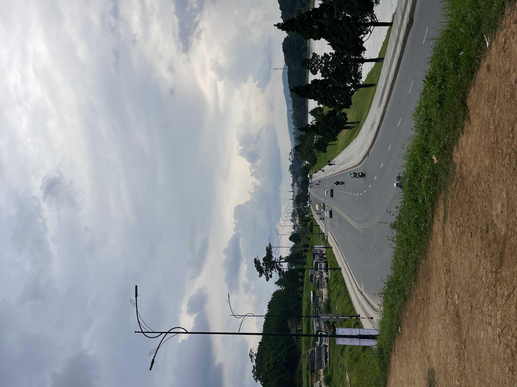 The image captures a scenic view of a road in Lâm Viên - Đà Lạt, Vietnam, under a cloudy sky. A dual-lane asphalt road curves to the right, bordered by a paved walkway and a line of tall, dark green cypress trees on the right. Beyond the trees, a body of water reflects the overcast sky. On the left side of the road, a grassy embankment slopes upwards, transitioning into a wooded area with more trees and some buildings in the distance. Several vehicles, including cars and motorcycles, are traveling on the road. A few pedestrians are visible on the walkway. In the foreground, the ground is a mixture of dry dirt and green grass. Streetlights are positioned along the left side of the road.
