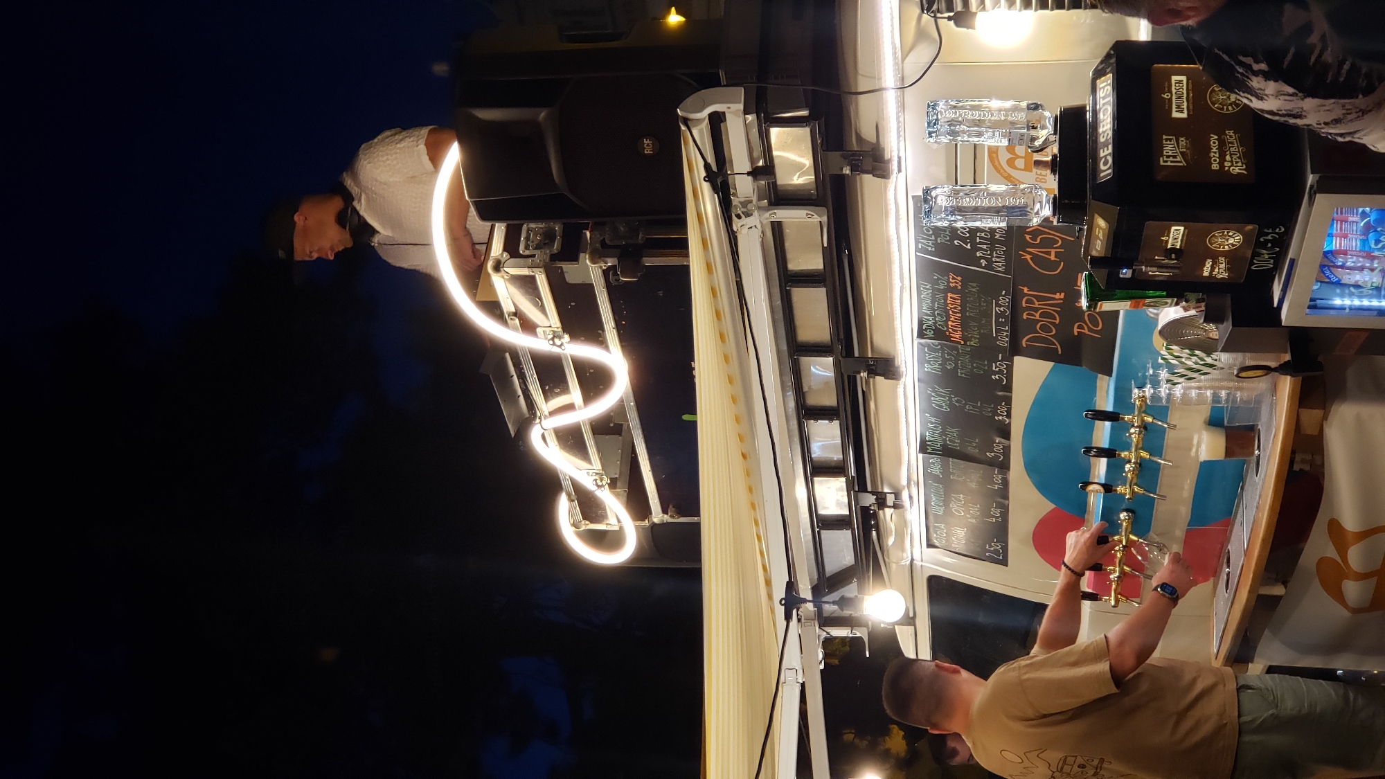 The image depicts an outdoor evening scene in the city of Prešov, Slovakia, possibly during a street festival or event. A man, likely a DJ, is visible on top of a vehicle, wearing a cap and headphones, and appears to be operating equipment. Below, a bar setup is present, with a man in a tan shirt standing behind the counter, interacting with beer taps. Several beer taps are mounted on the vehicle, with one glass of beer already poured. A menu board with various drink options and prices is displayed behind the taps. The scene is illuminated by decorative lights, including a neon-like light fixture above the DJ and spotlights. The overall atmosphere is lively and suggestive of a social gathering.