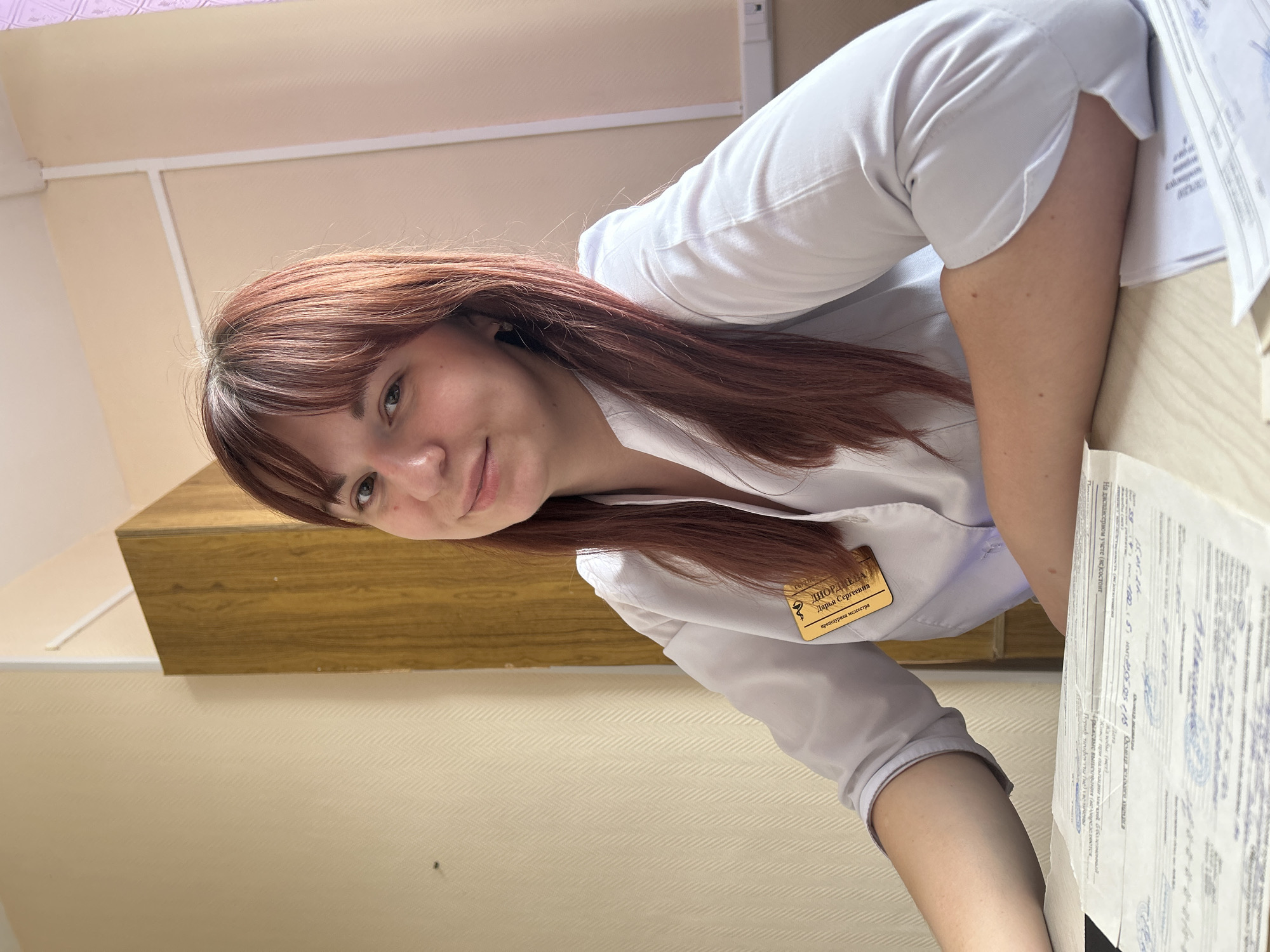 Here is a description of the image:
A young woman with reddish-brown hair and bangs, wearing a white medical coat, is positioned in front of what appears to be a wooden desk or counter. She is smiling and looking directly at the camera. A name tag, with some Russian text, is visible on her coat. There is a stack of papers to her right, positioned at an angle on the desk. The background includes a beige wall and a strip of what appears to be a cabinet.
Based on the OCR on the name tag, she may be a nurse in the city of Фоки, Russia.