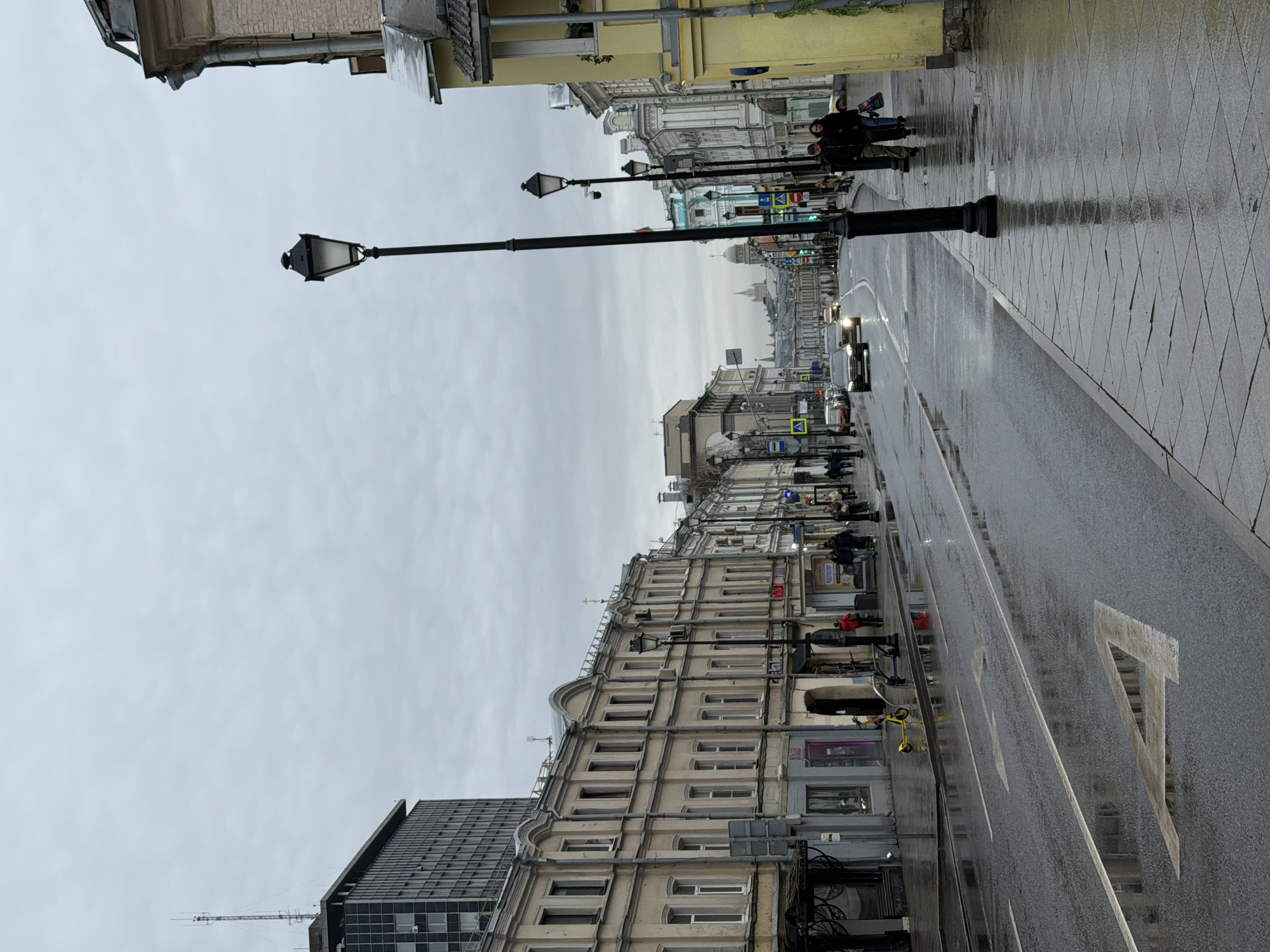 The image shows a street scene in Moscow, Russia, on a wet, overcast day. The camera is positioned at street level, looking down a long street lined with buildings on both sides.
The street is wet, with reflections of the buildings and sky visible on the pavement and road surface. White lines marking lanes and pedestrian crossings are visible on the road. A large, stylized "A" is painted on the rightmost lane of the road.
On the right side of the street, there is a paved sidewalk with a pattern of square tiles. A few people are walking on the sidewalk.
The buildings along the street are a mix of architectural styles, ranging from older, ornate structures to more modern buildings. Most of the buildings are several stories high, with rows of windows. Street lamps with a classic design are spaced along the street, lining the sidewalks.
In the distance, the street appears to converge, with buildings of varying heights. The sky is overcast, and the lighting is muted due to the weather conditions. There are cars driving on the road, and some pedestrian crossings are visible.
In the foreground, on the left side of the street, is a modern, taller building with a dark facade, contrasting with the older buildings.