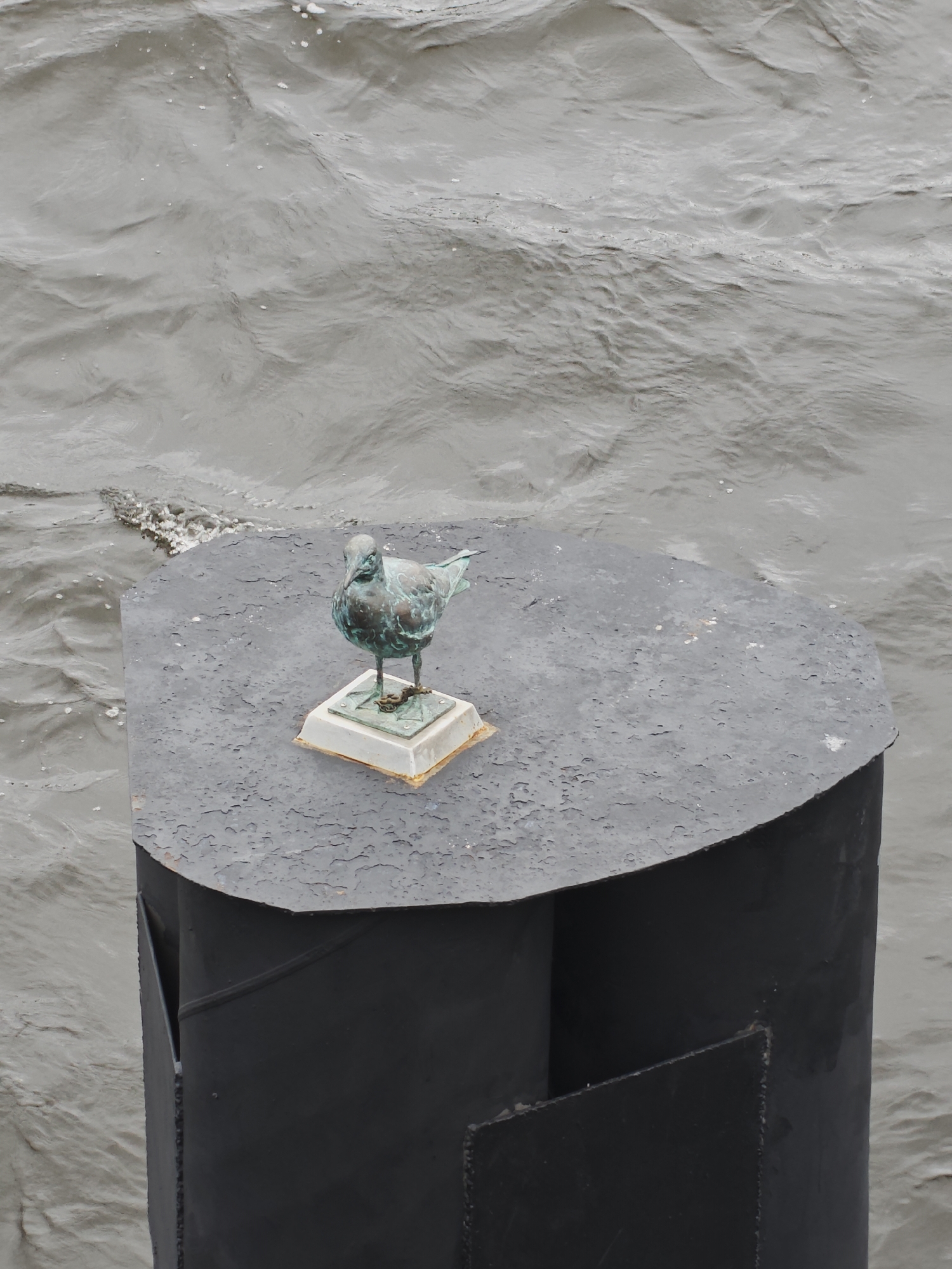A small, weathered bronze sculpture of a seagull stands on a black, metal surface on top of a black, cylindrical structure. The surface has a chipped, textured paint finish. The seagull is perched on a small, square, light-colored base, which is attached to a square metal plate. The base shows signs of corrosion and rust. The cylindrical structure has a curved top edge and appears to be made of metal. It is positioned on the water, with choppy, grey water surrounding it. In the background, the water shows ripples and some white foam or debris. This object is located at the Neva River embankment in Saint Petersburg, Russia, near the Summer Garden.