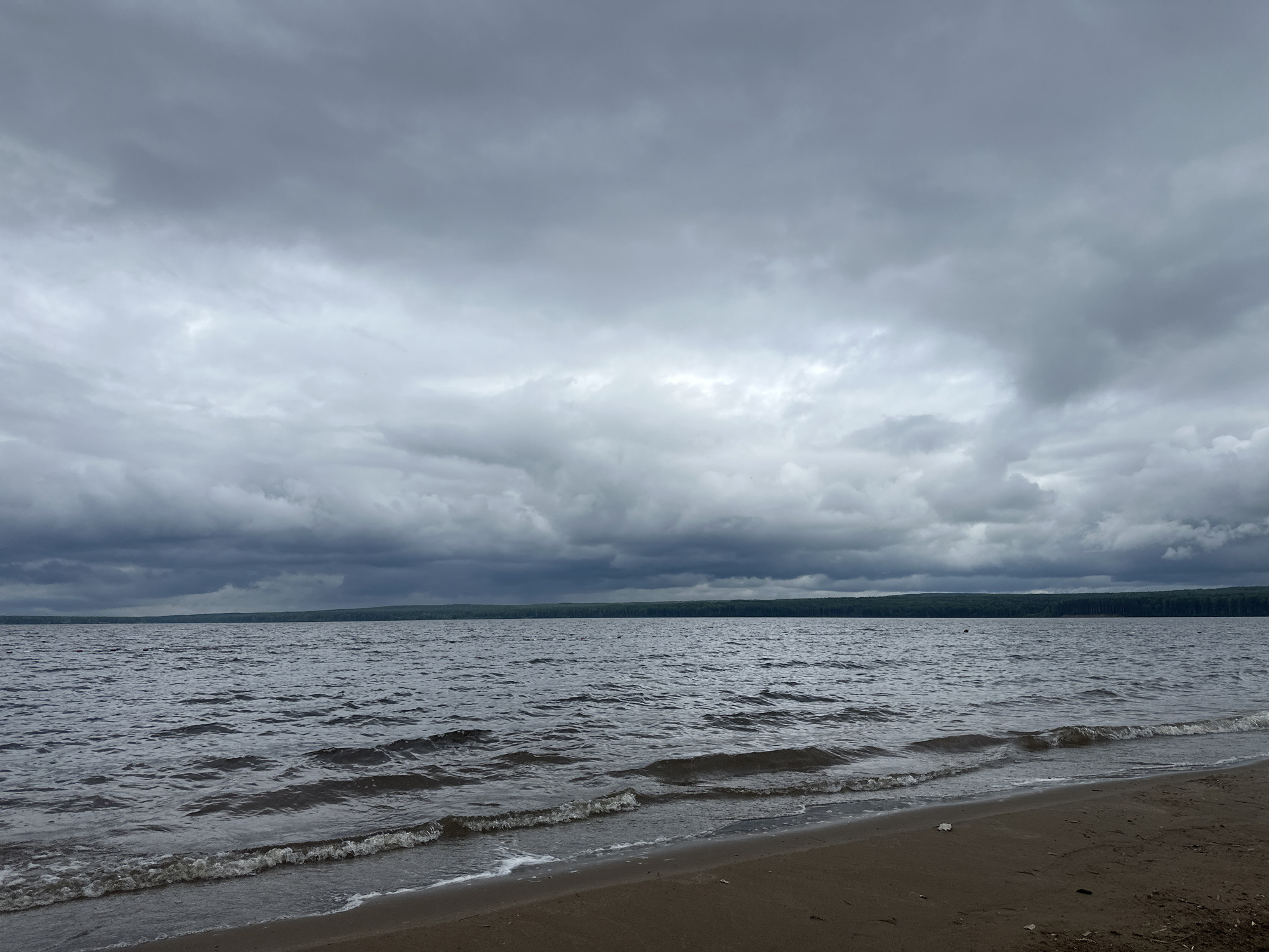 The image displays a wide-angle outdoor scene captured on a heavily overcast day, characteristic of the Votkinsk Reservoir shoreline in the city of Chaikovsky, Russia. The composition is horizontally divided into three primary sections: a sandy shoreline in the foreground, a body of water in the midground, and a cloud-filled sky above, with a distant tree line defining the horizon.
In the foreground, a strip of dark brown-grey sand forms a beach, sloping gently towards the water. Small, foamy waves are observed breaking at the water's edge, indicating gentle wave action. The sand appears wet where it meets the water.
The midground is dominated by a large body of dark, grey-blue water, exhibiting ripples and distinct wave patterns, suggesting wind presence. On the water, a small, dark, indistinct object is visible towards the right side, positioned in the mid-distance near the horizon.
The distant horizon is marked by a continuous, narrow band of dark green trees, indicating a forested shoreline.
The sky, occupying the upper portion of the image, is entirely covered by a thick layer of grey, multi-layered stratus or nimbostratus clouds. These clouds range in tone from lighter grey in the center to darker, denser grey at the top and along the tree line, without any discernible breaks or direct sunlight.
No individuals, animals, or man-made structures are prominently featured, except for the aforementioned small, dark object on the water. The scene conveys a sense of quiet, natural landscape under a cloudy sky.