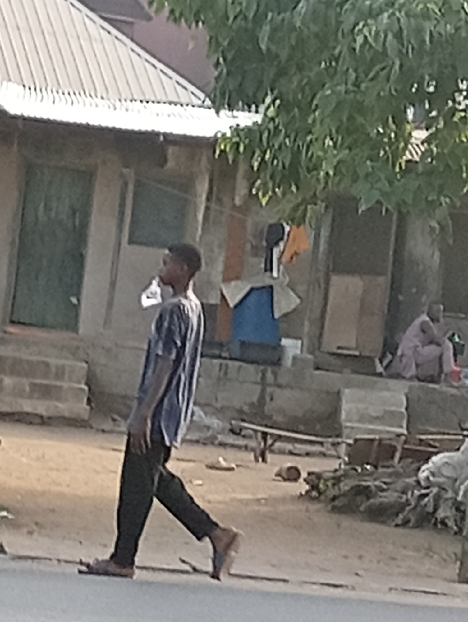 The image depicts an outdoor street scene in Adankolo, Nigeria, during what appears to be either early morning or late afternoon, characterized by soft, warm lighting.
In the foreground, a young man walks from left to right, captured in profile. He wears a striped t-shirt, dark trousers, and light-colored sandals. His head is turned slightly forward, suggesting he is looking ahead.
The background features several simple, single-story buildings with corrugated metal roofs. On the left, one building has a green wooden door and is approached by a set of concrete steps. To its right, another structure displays laundry hanging to dry, including an orange garment, a white piece of clothing, and some darker fabric, situated near a blue plastic container and what appear to be wooden planks or cardboard sheets. Further into the mid-ground on the right, a person is seated on a raised concrete step or platform, dressed in a light-colored, long robe-like garment and possibly red shoes.
The ground throughout the scene is unpaved, appearing dusty and uneven, with scattered debris and a few wooden objects, including a log, visible. A large tree with dense green foliage partially overhangs the upper right side of the image, providing shade. The overall setting suggests a residential area with modest housing. There is no visible text in the image.
