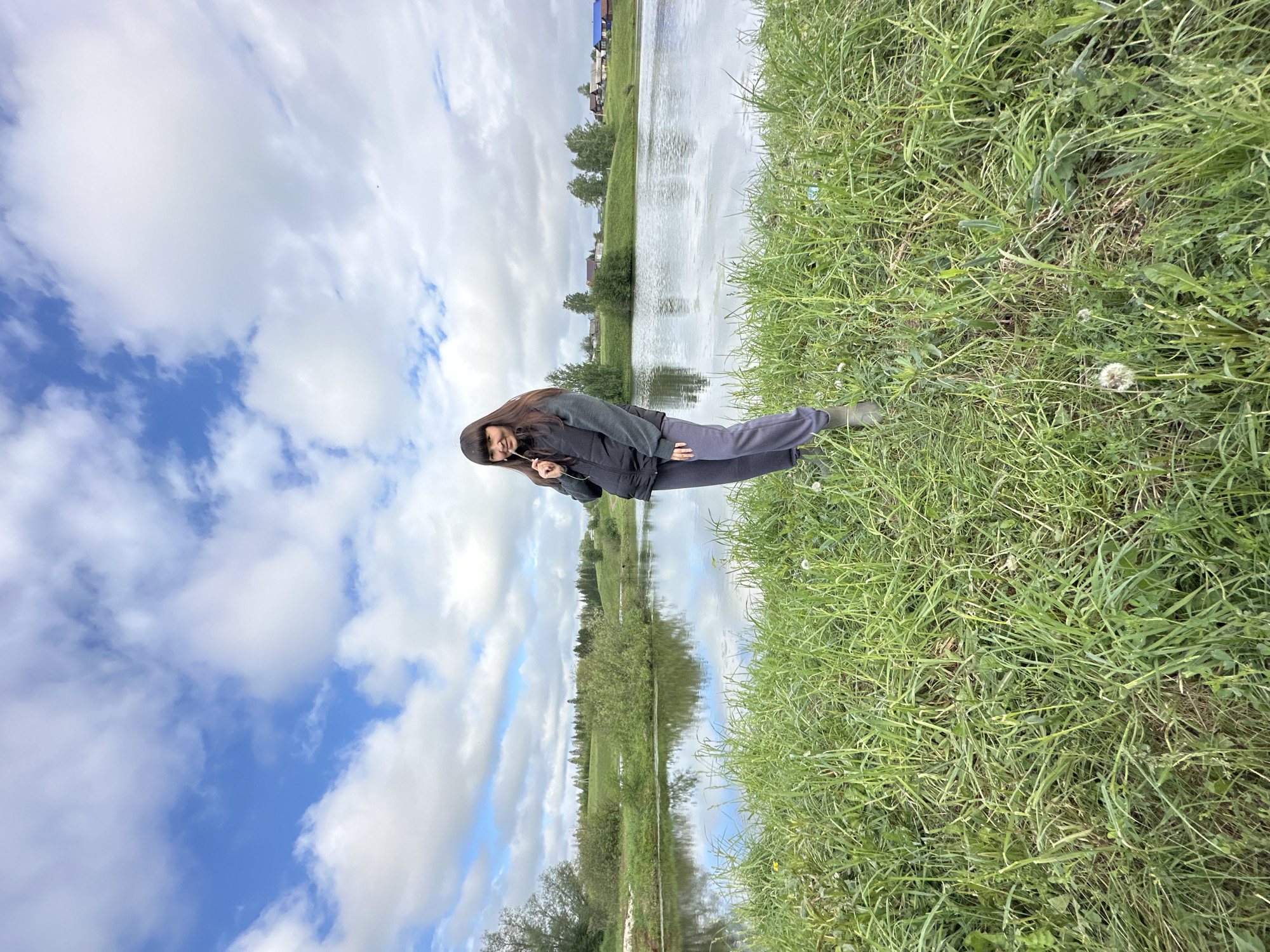 A photograph taken in Среднее Кечево, Russia, depicts a woman standing in a grassy area next to a body of water, presumably a pond or lake. The woman has long, dark hair, and is wearing a dark jacket and pants, and what appear to be boots. She is holding a thin, white object, possibly a stick or stem, near her face. The body of water reflects the sky, which is filled with clouds and patches of blue. Across the water, there is visible vegetation and a few buildings that define the location. The immediate foreground consists of tall green grass.
