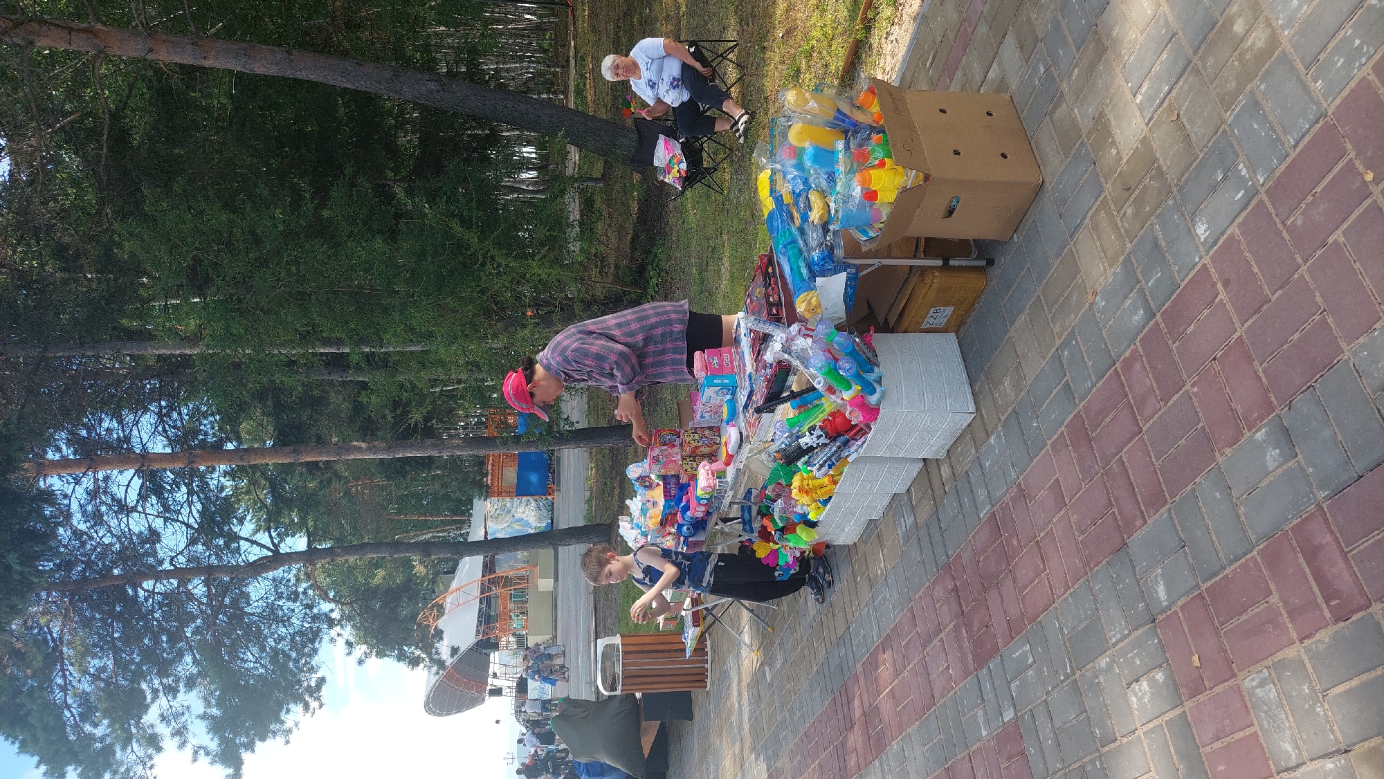 The image depicts an outdoor scene, likely a park or waterfront area, with a stall selling toys. In the foreground, a young boy wearing a blue and black striped tank top and dark pants is looking at a toy in his hands. Beside him, a woman wearing a red cap and a plaid shirt with black shorts stands behind a table laden with various colorful toys, including toy guns, water pistols, and other small play items. In the background, an older woman is seated in a folding chair, observing the scene. The ground is paved with red and gray interlocking bricks. Further back, a stage-like structure is visible, suggesting a public event or recreational area. Trees form a dense backdrop, and the sky is visible above. The setting appears to be a sunny day. The image does not contain any specific landmarks or visual cues that would allow for definitive identification of Komsomolsk-on-Amur, Russia.