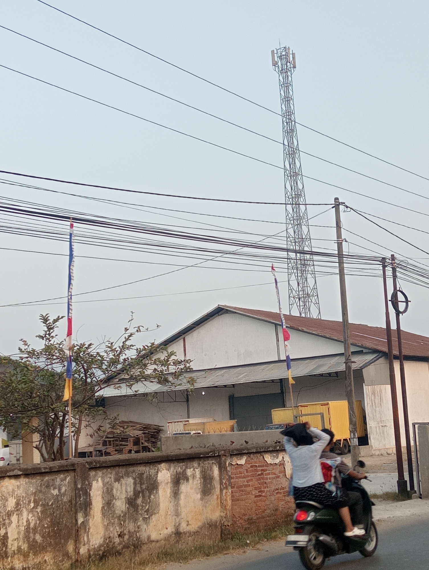 The image shows a street scene in Pringsewu, Indonesia. A cellular tower is visible in the background, with multiple power lines crisscrossing the frame. In the middle ground, there is a white building with a rusty roof and a stack of wooden planks to its left. A yellow truck is parked in front of the building. In the foreground, two people are riding a scooter on a paved road. The person in the back is wearing a light-colored shirt and dark patterned shorts, with their hands raised to their head. The person in front is wearing darker clothing. A concrete wall with peeling paint and some brickwork runs along the side of the road. There are flags on poles in front of the building. The sky is overcast.