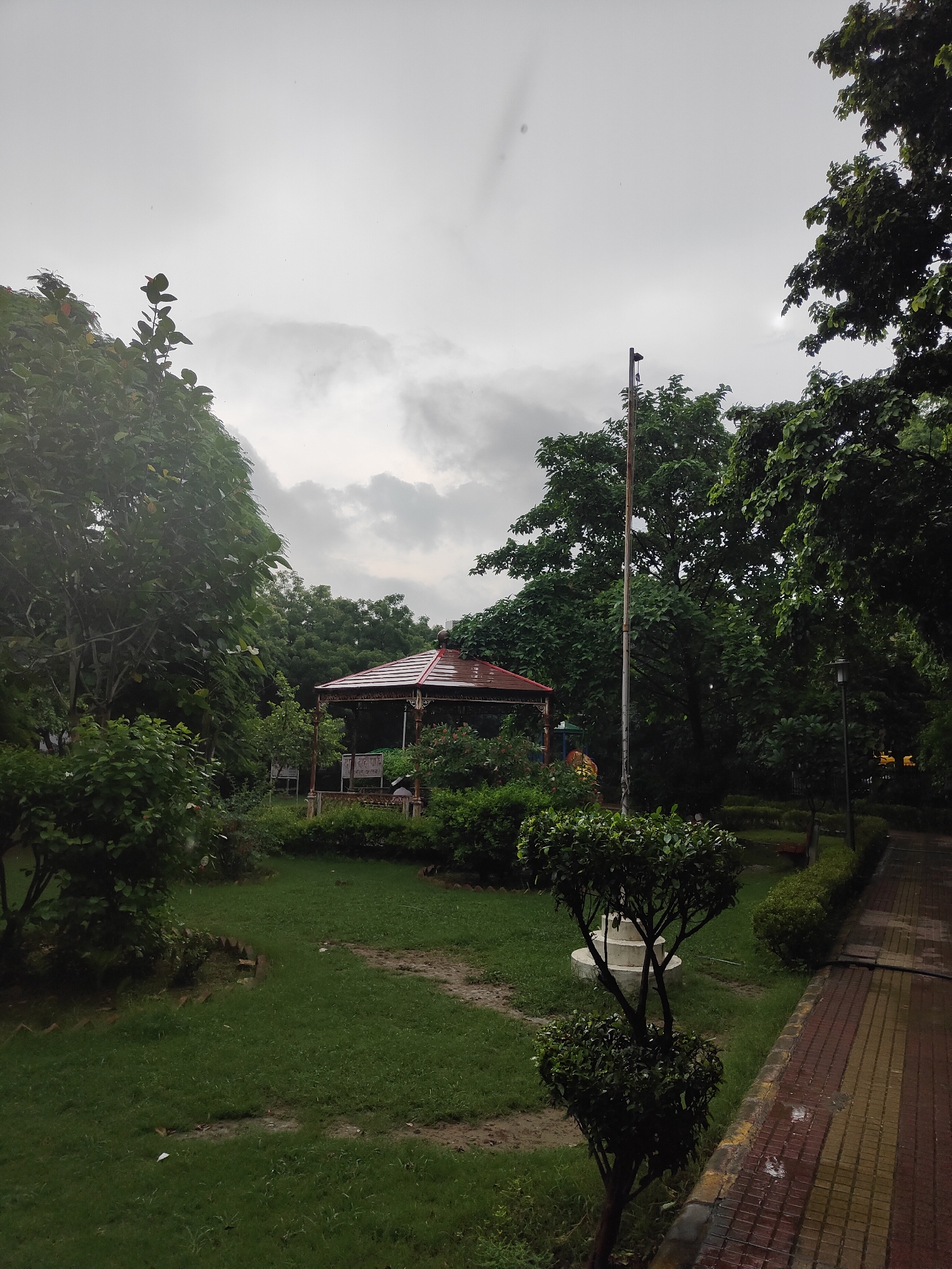 The image depicts a park in Gurgaon, India, on a cloudy day, possibly after rain given the wet pavement. In the center of the scene is a gazebo with a red, angular roof and white supports, surrounded by lush greenery. A flag pole stands tall to the right of the gazebo, with a lamp post visible further down the paved walkway. The walkway itself is made of red and yellow bricks and shows signs of being wet. The park features well-maintained grass areas, manicured bushes, and mature trees that create a dense canopy in places. The overall atmosphere is serene and green, typical of a public park. There are no people visible in the image.