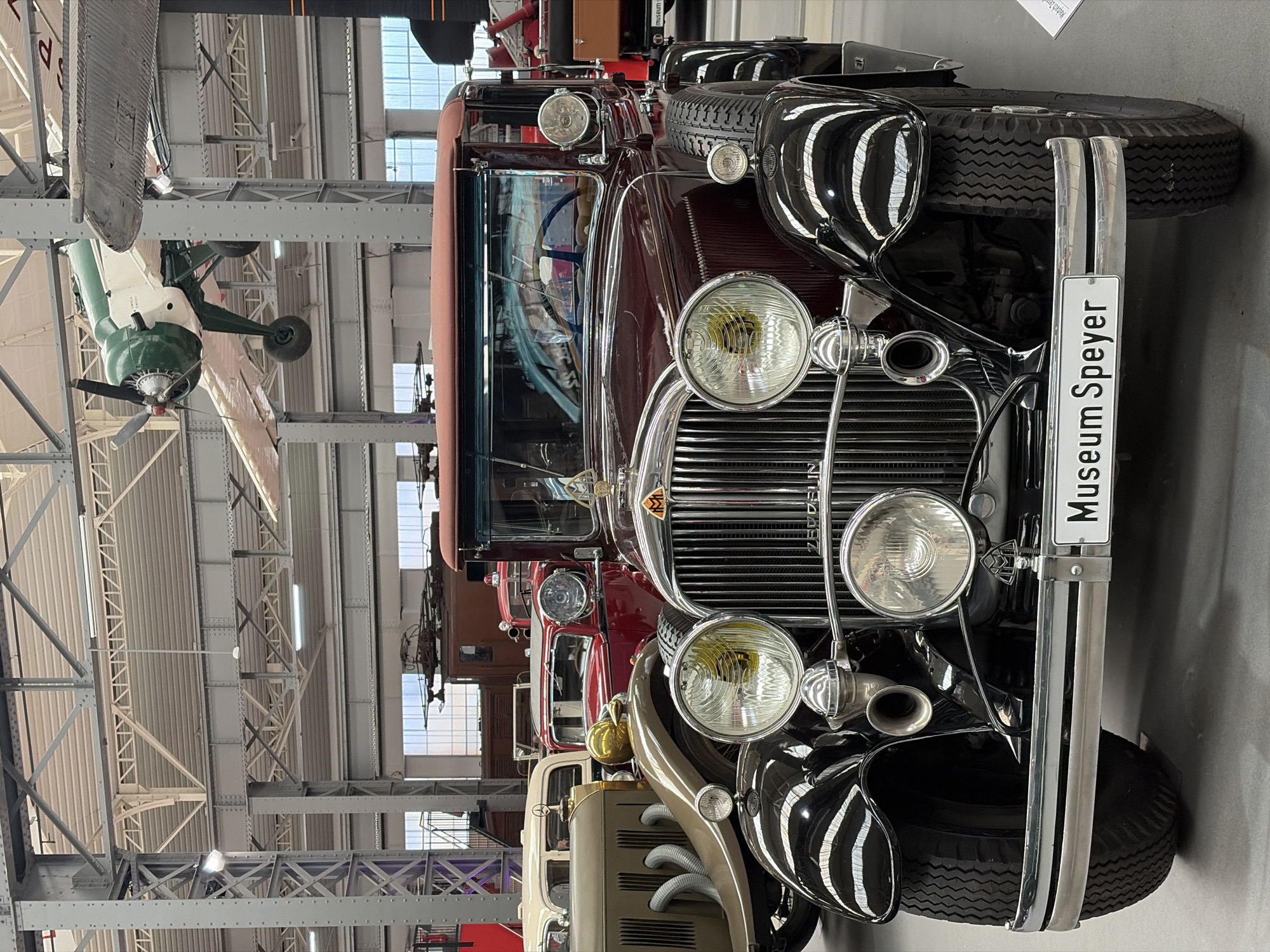 This image shows the front of a dark red vintage automobile displayed in a museum. The car has a prominent vertical grille with chrome accents, two large circular headlights on either side of the grille, and a smaller headlight positioned below the grille. The vehicle features a visible emblem on the grille and the word "ZEPPELIN" inscribed above it. The car is fitted with large, black tires. In front of the car, a white license plate reads "Museum Speyer". The car appears to be indoors, within a large hall with a high ceiling supported by metal beams. Other vintage vehicles, including a lighter-colored car and parts of what appear to be trains, are visible in the background, along with an airplane suspended from the ceiling. The lighting in the museum is a mix of natural light from windows and artificial lighting from fixtures on the ceiling. There are no people visible in the image. The overall scene suggests a display within the Speyer technical museum.