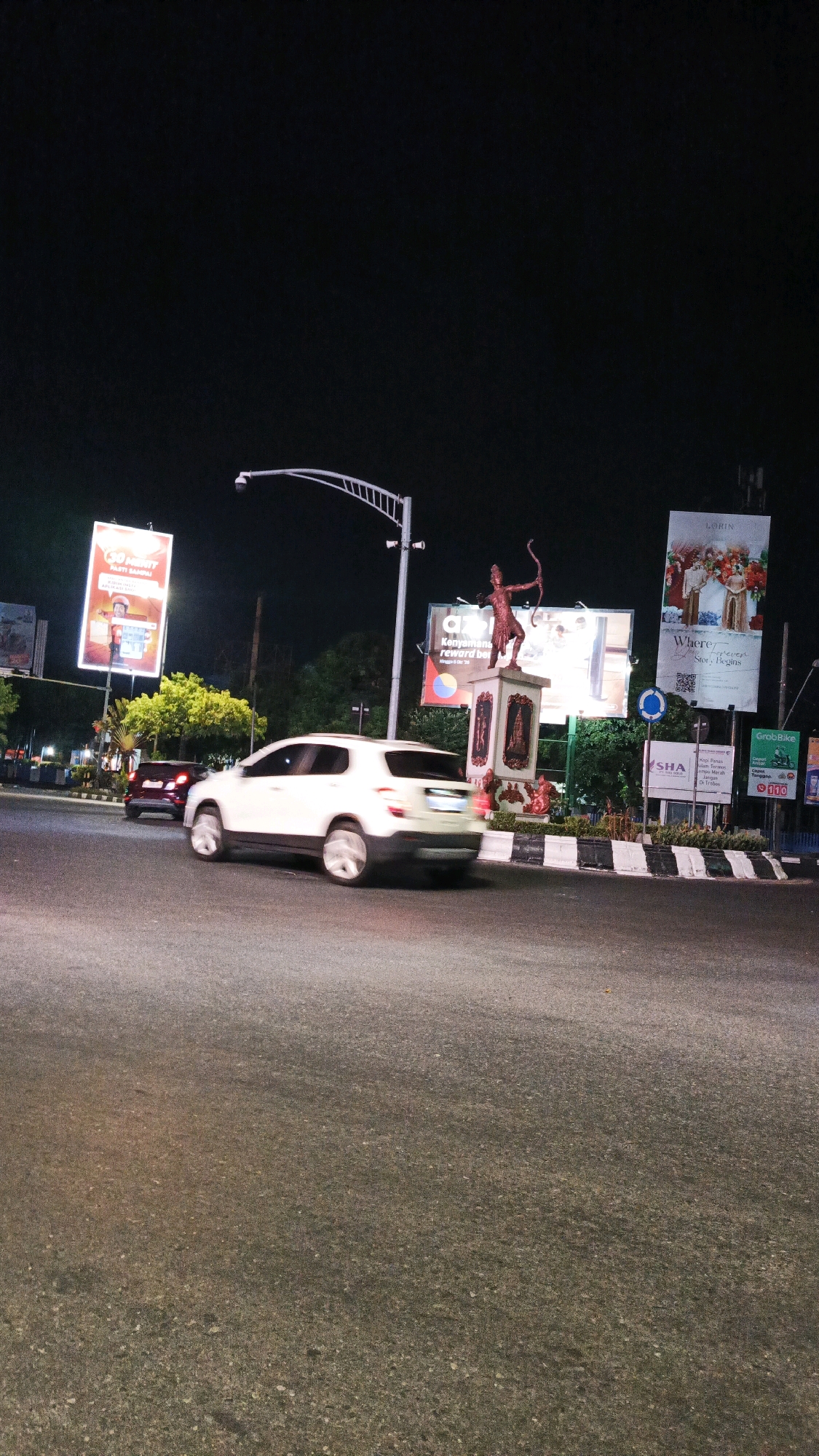 This image depicts a night scene at a traffic roundabout in Surakarta, Indonesia. A white SUV is captured in motion, blurred due to the long exposure, as it navigates the roundabout. In the background, a statue of a figure holding a bow and arrow stands prominently on a pedestal, illuminated by spotlights. Several billboards with advertisements are visible around the roundabout, including one with text in Indonesian and an image of a person in orange attire, and another with an image of a couple in wedding attire. The environment is urban, with streetlights and trees contributing to the nocturnal ambiance. The road surface appears to be asphalt.