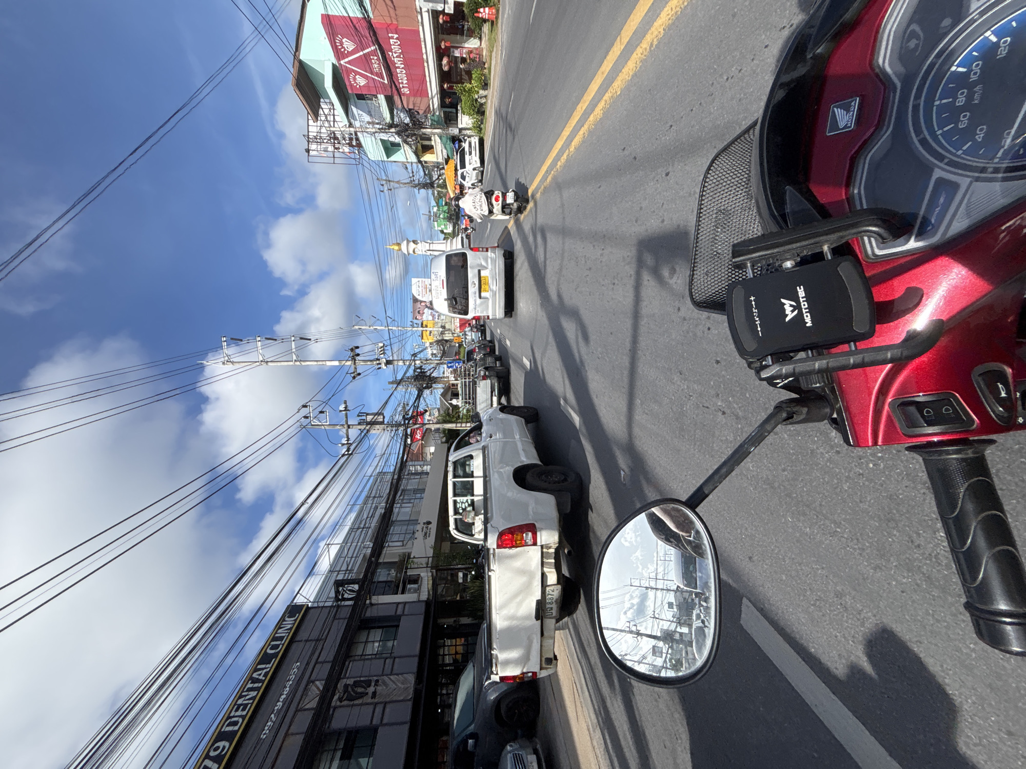 The image depicts a street scene in Chalong, Thailand, taken from the perspective of someone riding a red motorcycle. The rider's handlebars and part of the motorcycle's dashboard, including a phone mount with the brand "MOTOTEC" visible, are in the foreground. The road is paved with asphalt and marked with yellow lines. A white pickup truck is in the left lane, and a silver van is in the right lane. Further down the road, a person on a scooter wearing a white jacket with the text "CREATIVE ATHLETICS" is visible. Buildings line the right side of the street, with various signs and advertisements displayed. Overhead, a complex network of electrical wires crisscrosses the sky, which is blue with scattered clouds. The reflection in the motorcycle's side mirror shows other vehicles and buildings. The overall environment suggests a typical day on a busy street in a Thai city.