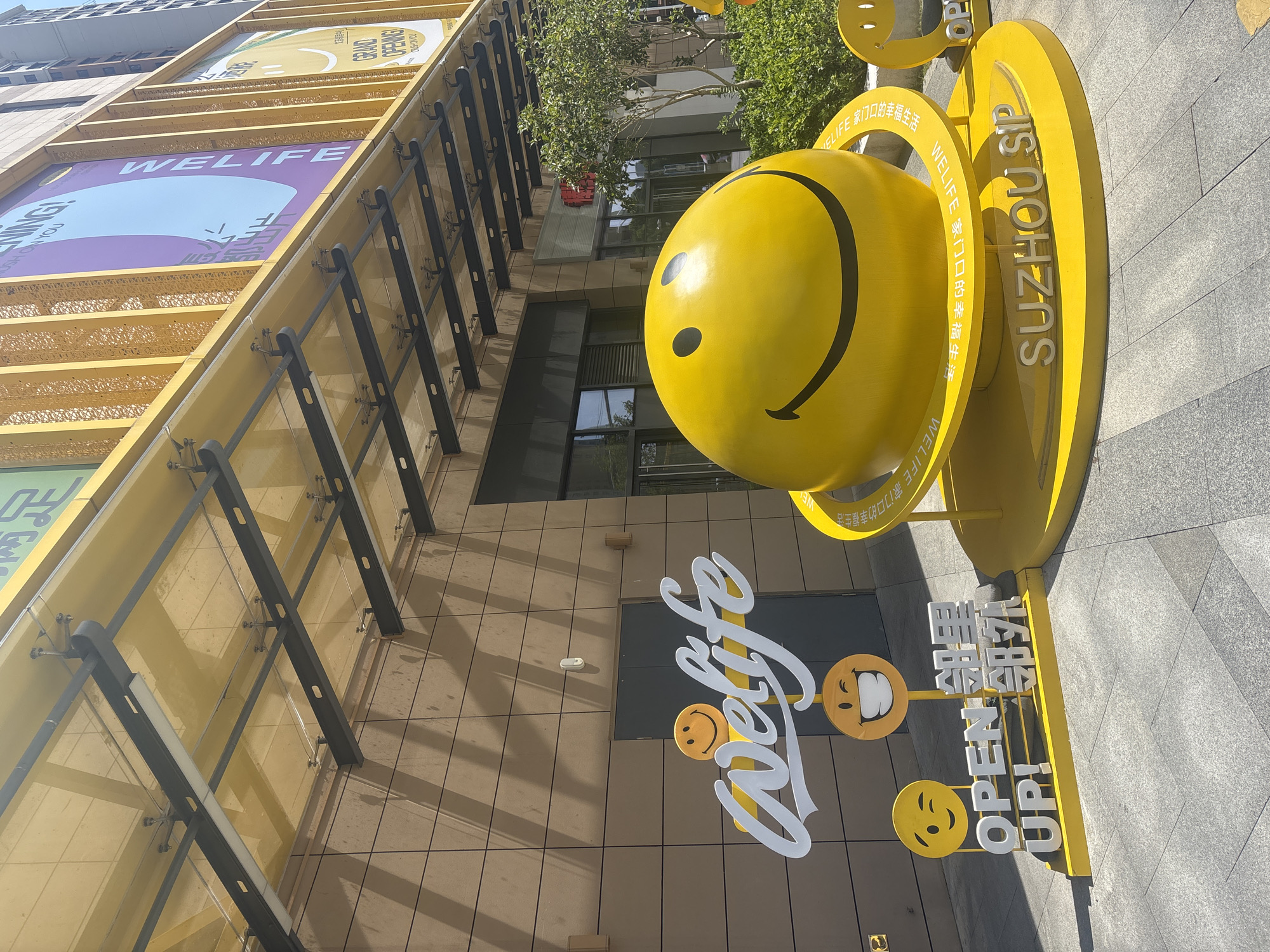 A bright yellow smiley face sculpture is prominently displayed on a circular yellow platform in front of a modern building with a beige tiled facade. The sculpture is a large sphere with a classic smiley face painted on it, and it is surrounded by a yellow ring with text in Chinese. In front of the sculpture, there is a white "welife" logo with a small smiley face above it and a winking smiley face below. To the left of the "welife" logo, a yellow stand displays the English text "OPEN UP!" alongside Chinese characters. The ground is paved with grey rectangular tiles. The building has a yellow awning extending over the entrance, supported by dark metal beams. Large banners with text and imagery, including a purple banner with "WELIFE" and a blue circle, are visible on the upper part of the building. The scene is set in the Suzhou Industrial Park, China, as indicated by the text on the platform. The lighting suggests a sunny day, casting shadows on the ground. There are no people visible in the immediate foreground.