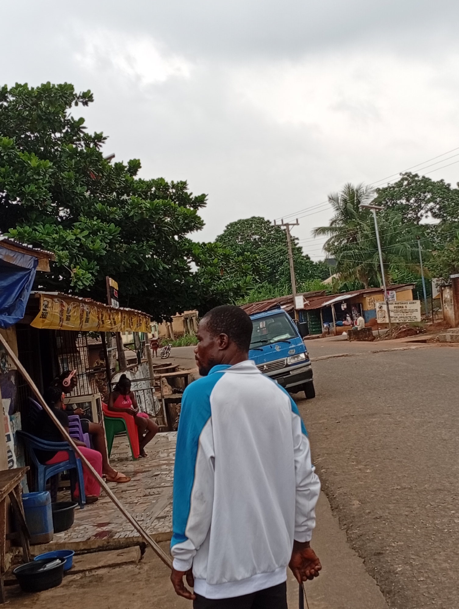 A medium-shot, eye-level, outdoor, day time, slightly overcast, vertical, medium saturation, full focus, Nigerian street scene shows a man in the foreground from the shoulders down, wearing a white and blue jacket, facing away from the camera and looking to his left. He appears to be walking. To his left, in the mid-ground, are three women, two of whom are seated on chairs, with one on the far left wearing headphones, and the woman in the center wearing pink clothing. A blue van is parked on the right side of the street in the mid-ground, and a street sign is visible on a pole in the background on the right. The environment is a street with shops and trees. The street scene is located in Nsugbe, Nigeria.