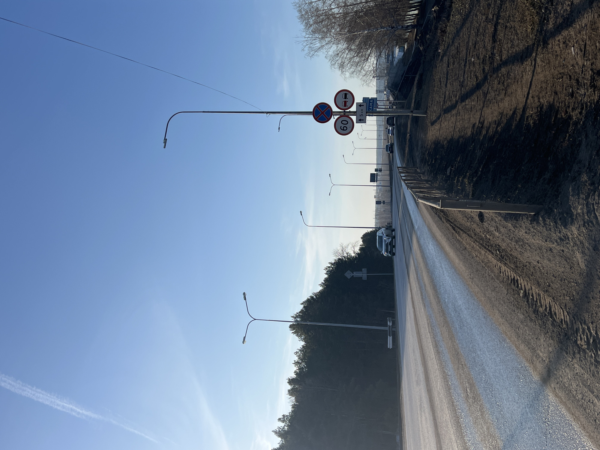 The image depicts a road in Chaikovsky, Russia, recognizable by the "KAMA 5 km" sign. The view is looking down the road, which is wet and reflects sunlight. On the right side of the road is a dirt bank with vegetation. The sky is clear and blue. Streetlights line the road. Traffic signs are visible, including one indicating a speed limit of 60 km/h and a no-passing zone. A few vehicles are on the road. The image appears to be taken on a sunny day.