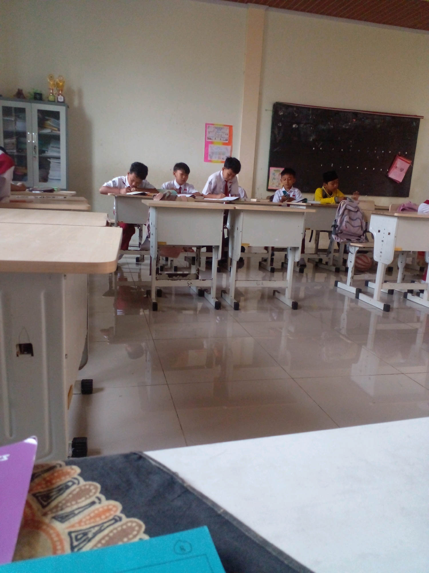In a classroom setting in Banyuputih, Indonesia, several students are seated at desks, engaged in schoolwork. The room features a blackboard on the right side of the wall, adorned with various papers and what appears to be educational materials. A cabinet with glass doors stands to the left, displaying books and trophies on its upper shelf. The floor is tiled and reflective, showing the overhead lighting and the arrangement of student desks and chairs. The students, dressed in school uniforms, are focused on their books, with some writing and others reading. The overall atmosphere suggests a typical school day.