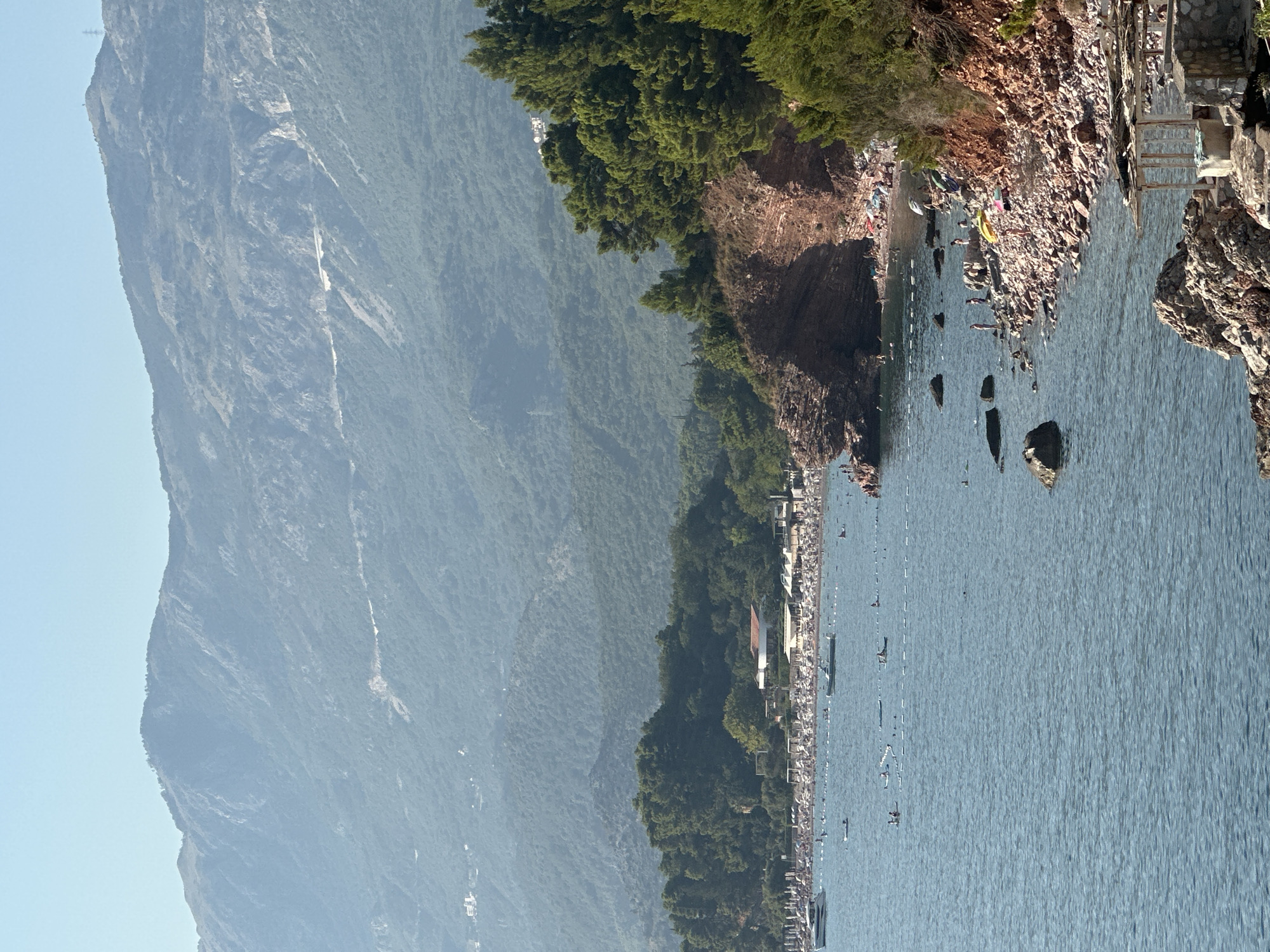 This is a vertical, eye-level shot of a rocky coastline with a beach and mountains in the background, likely taken in Sveti Stefan, Montenegro. The foreground is dominated by the blue water of the Adriatic Sea, with several rocks scattered along the shoreline. The shoreline itself is a mix of sand and pebbles, with a rocky cliff face covered in lush green trees.
A beach is visible in the mid-ground, lined with white umbrellas and lounge chairs, indicating a popular tourist destination. Numerous people are scattered on the beach and in the water, some swimming, sunbathing, or engaging in water sports like kayaking and paddleboarding. Several boats, including a white yacht and smaller vessels, are moored in the bay.
In the background, dense green mountains rise up, their slopes covered in trees and dotted with hints of human habitation. The sky is a clear, pale blue, with a slight haze obscuring the distant peaks. A simple wooden structure, possibly a beach shelter or changing room, is visible on the rocky shore in the lower right corner.