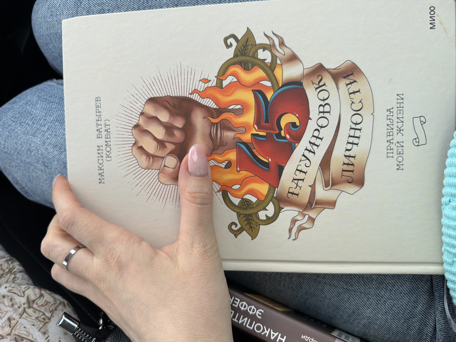 The image presents a close-up, overhead view of an individual's lap, located in the city of Yus'ki, Russia. A light-skinned left hand, wearing a thin silver-colored ring on the ring finger and exhibiting clear-to-light-pink ombré manicured nails, is positioned on the right side of a cream-colored hardcover book. The book's cover features Russian text, including "МАКСИМ БАТЫРЕВ (КОМБАТ)" at the top, and "45 ТАТУИРОВОК ЛИЧНОСТИ" with "ПРАВИЛА МОЕЙ ЖИЗНИ" at the bottom, along with a "МИФ" publisher logo. The central illustration on the cover depicts a stylized clenched fist emanating light rays, situated above the large red numbers "45" surrounded by orange flames, all overlaid on a beige banner. The book rests upon blue denim jeans. To the left, partially obscured, is a second book with a dark brown spine, visible with the text "НАКОПИТЕЛЬНЫЙ ЭФФЕКТ". In the upper left corner, a dark fabric with a silver-colored zipper pull is visible. A light blue, ribbed knitted fabric is partially discernible at the bottom edge of the frame.