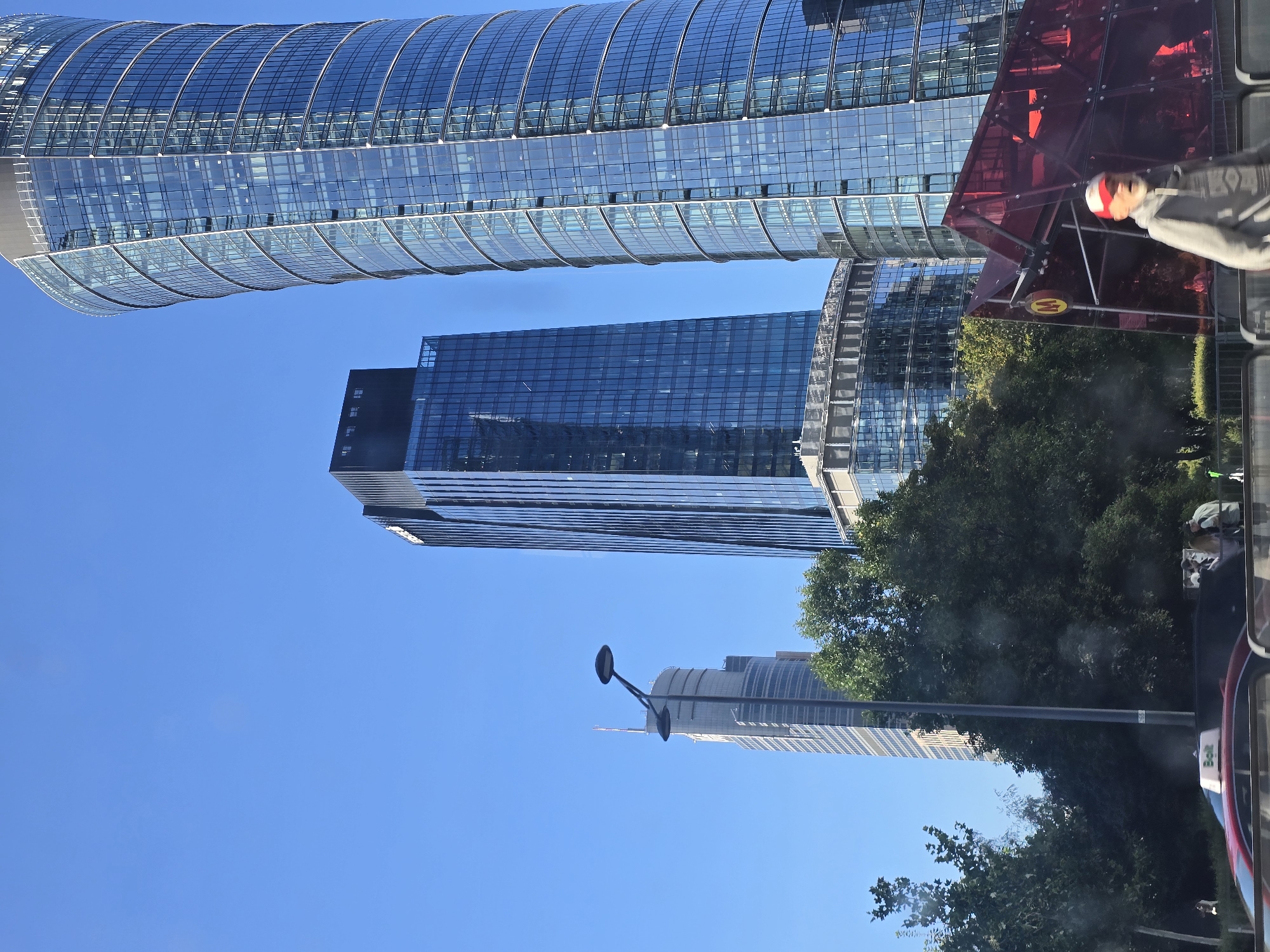 The image depicts a street-level view of a modern urban environment in Warsaw, Poland, dominated by several contemporary skyscrapers. On the right, a tall, cylindrical building with a blue glass facade curves upward, its many windows reflecting the clear blue sky. To its left, a narrower, rectangular skyscraper with a dark glass facade rises, also reflecting the sky. Further in the background, a lighter-colored, slender skyscraper is visible, with a lamppost in the foreground partially obscuring it. Lush green trees line the street, interspersed between the buildings and the foreground elements. In the immediate foreground, a red structure with a geometric design, possibly a bus shelter or an architectural feature, is visible. A man wearing a red and white cap and a grey hoodie is walking past the red structure. To the left of the red structure, a "M" symbol, indicating a metro station, is mounted on a circular sign. A red car with a "Bolt" sign on its roof is parked on the street. The overall scene suggests a bustling city center with modern architecture and public transportation infrastructure.