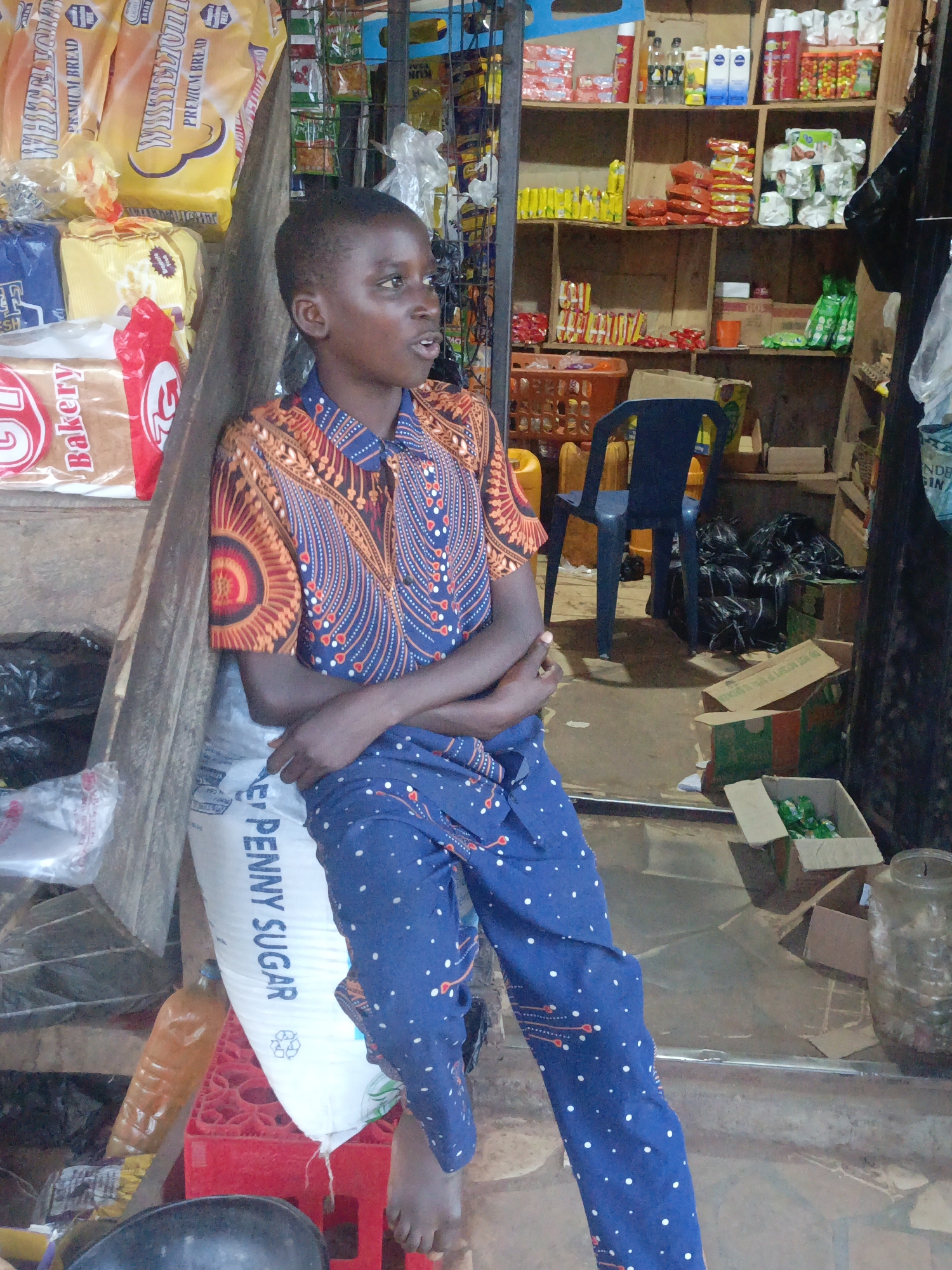 A person stands leaning against a bag of "PENNY SUGAR" inside a cluttered store. The individual is wearing a patterned shirt in shades of blue and orange, paired with blue pants dotted with white spots. Their arms are crossed.
The environment is a shop containing assorted goods. Stacks of wrapped bread are visible to the left, while shelves behind the subject hold various items, possibly food or household supplies. A dark blue plastic chair is partially visible in the background. Boxes and containers scattered on the floor add to the cluttered appearance.
The location is Akwanga, Nigeria, based on the presence of the "PENNY SUGAR" bag, which is commonly found in the region.