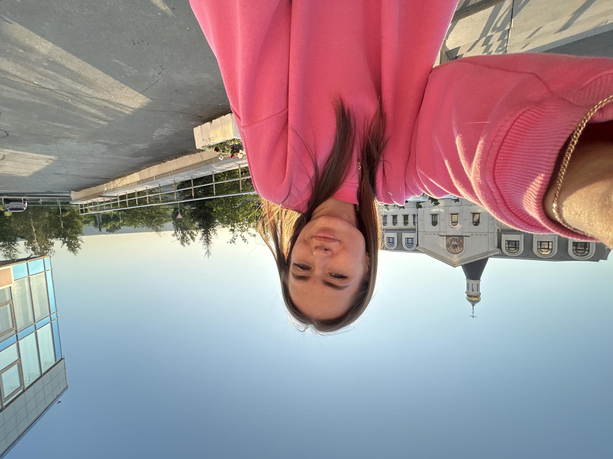A young woman with shoulder-length brown hair is taking a selfie outdoors during the daytime. She is wearing a bright pink oversized sweatshirt and a gold chain necklace. The background features a large white building with a gold dome and cross, resembling a church or monastery. To the right, a modern building with a blue glass facade is visible. The scene appears to be in an urban or suburban setting with trees and a paved walkway. The sky is clear and blue, and the sun is casting shadows on the ground.