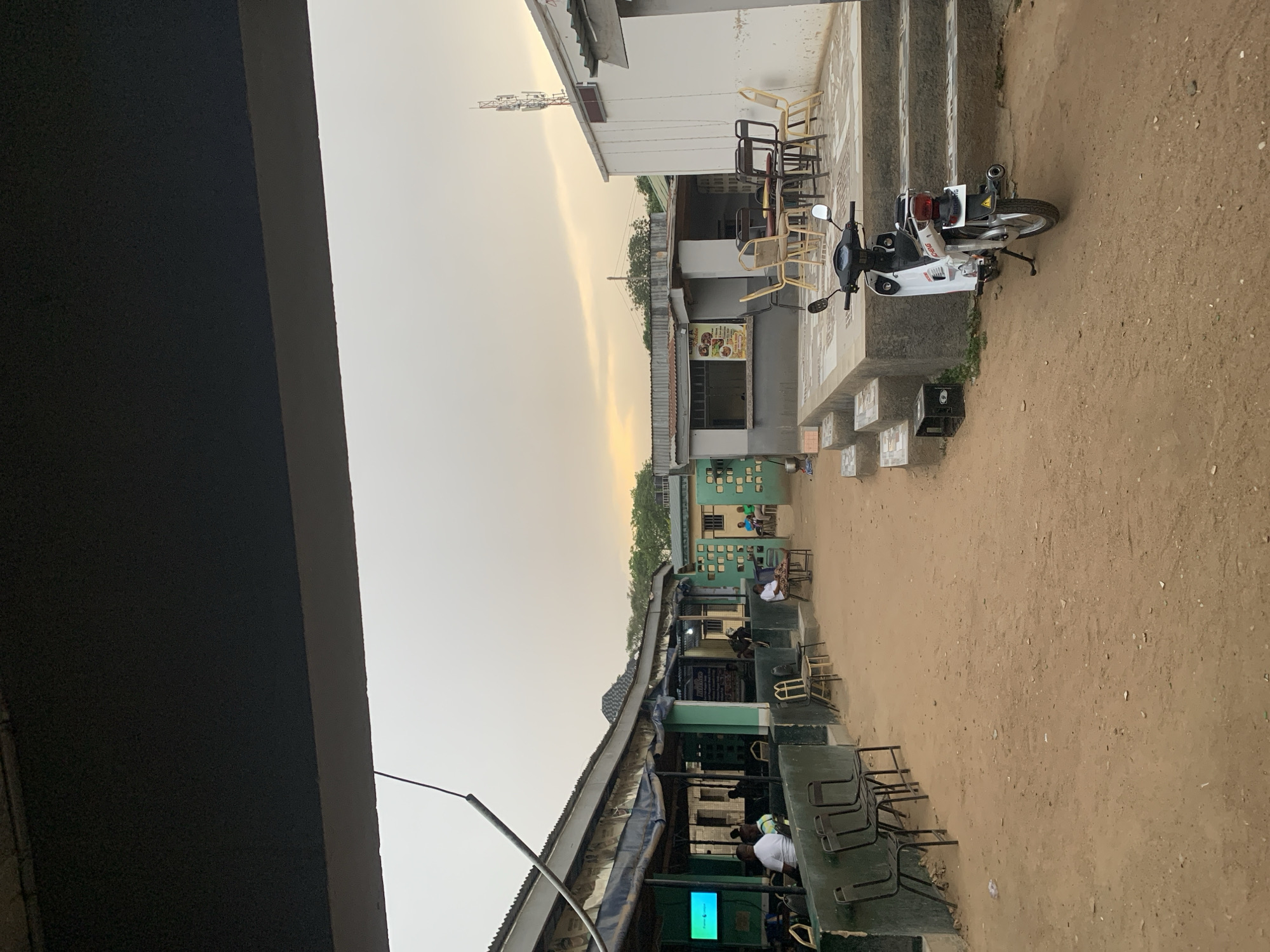 The image depicts an outdoor scene in Kabong, Nigeria. Buildings line a sandy area. People are sitting and standing near the buildings. A motorcycle is parked next to a building with steps. There are chairs placed on a patio. A communication tower rises in the distance. The sky is overcast.