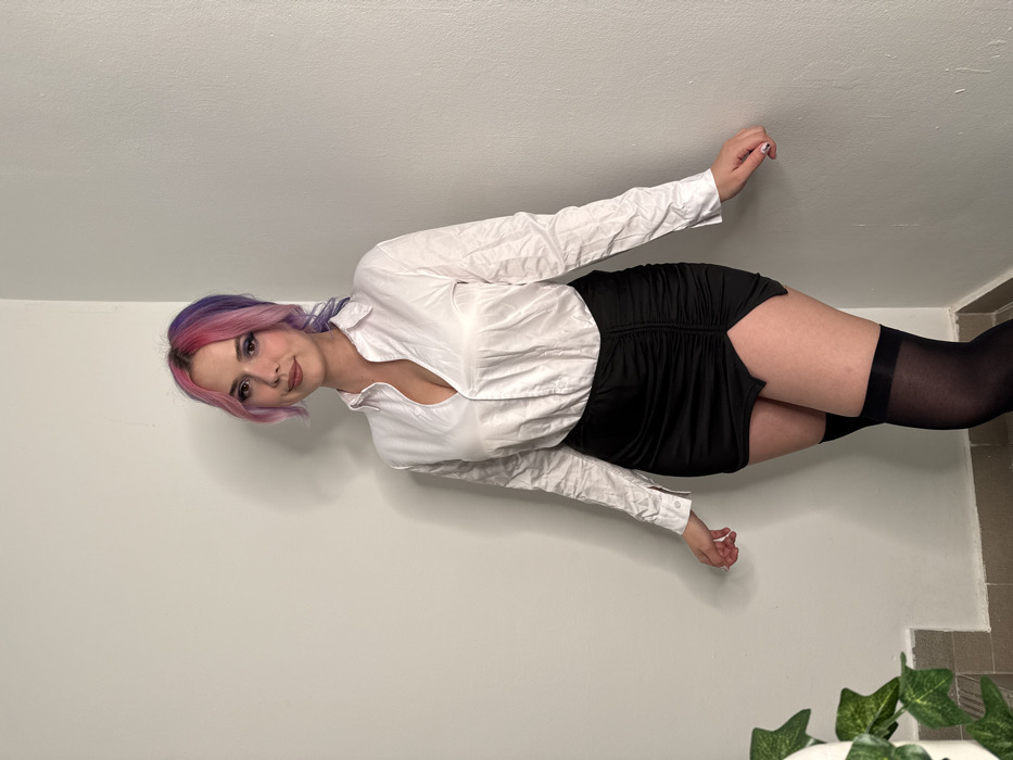 An image depicts a single young adult female standing indoors in what appears to be a corner of a room. The individual is positioned centrally, facing the camera directly, and exhibits a slight smile. Her hair is split-dyed, with vibrant pink on the viewer's left side and purple on the viewer's right, styled with a side part. She wears a white long-sleeved button-up shirt, deeply unbuttoned at the collar, revealing cleavage. Below the shirt, she wears a black mini skirt with front ruching and an asymmetrical hemline. Her legs are covered by black thigh-high stockings. The background consists of plain, light-colored walls forming an interior corner, with a floor visible at the bottom. A small green leafy plant is partially visible in the bottom-left corner of the frame, situated next to the wall. The specific location within the City of Zagreb, Croatia, cannot be determined from the visual evidence present in the image.