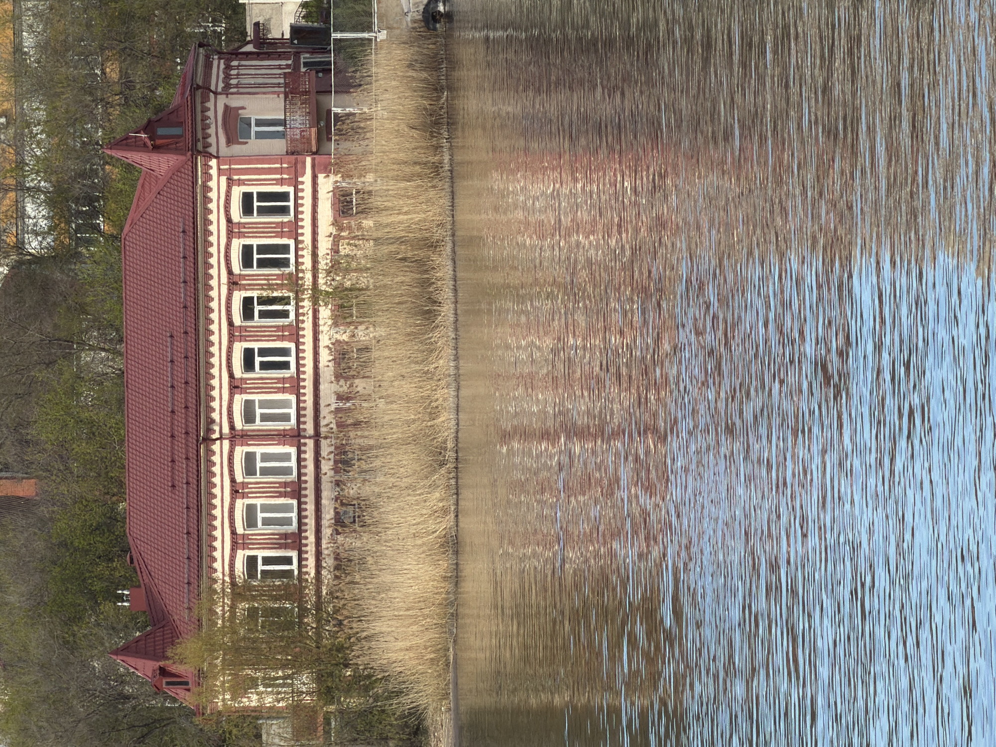 A four-story building with a red roof and light-colored walls with white window frames stands next to a body of water, likely a river or lake. Tall, dry grass or reeds grow between the building and the water. The water reflects the building and sky. The photo was taken in Kamyshin, Russia.