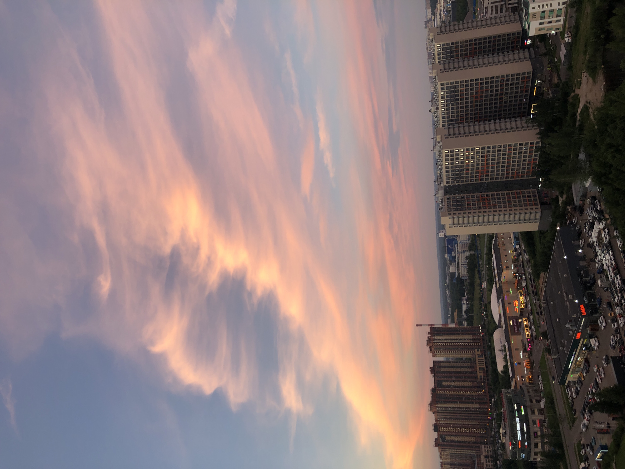 This is a high-angle, vertical, full shot of a city landscape at dusk. The sky is filled with wispy clouds tinged with pink and orange hues, contrasting with the blue sky above. In the foreground, several multi-story apartment buildings dominate the scene, with numerous windows visible, some illuminated. Below the buildings, a busy commercial area is apparent, featuring parking lots filled with cars and various retail establishments, including one with a "LADA" sign. Trees and some smaller structures are scattered throughout the urban environment. In the distance, more buildings and a hint of industrial infrastructure can be seen. The overall impression is that of a developing urban area during the evening hours, with a striking sky.
