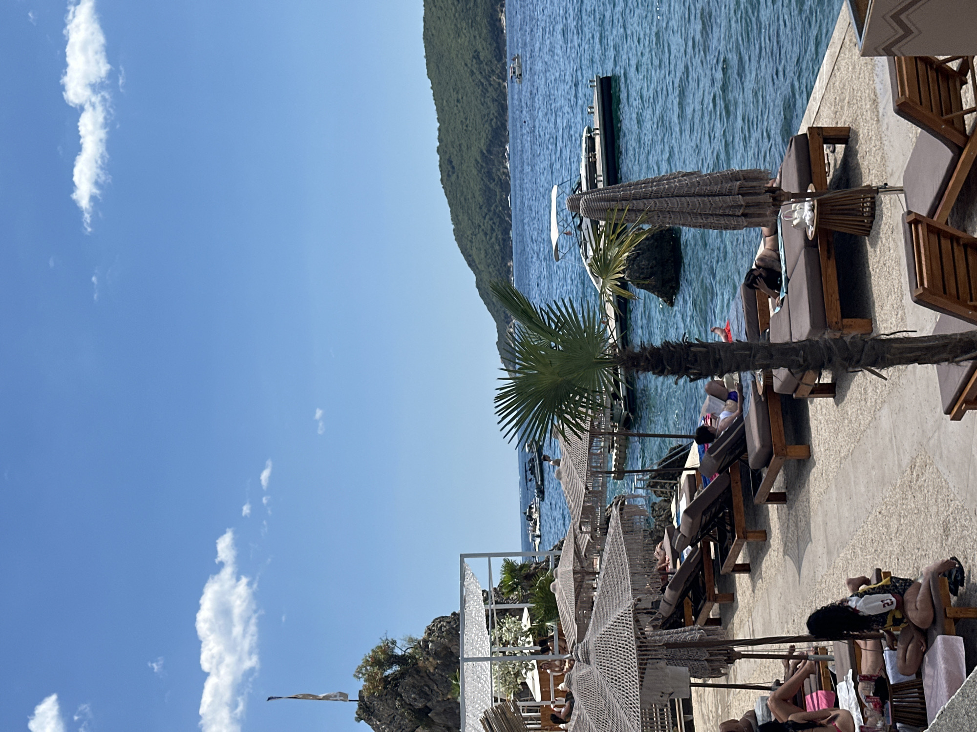 The image depicts a sun-drenched beach scene in Budva, Montenegro, likely at a resort or private beach club. In the foreground, several lounge chairs are arranged on a paved area adjacent to the turquoise water. Some chairs are occupied by individuals sunbathing, while others are empty. Woven sun umbrellas provide shade over some of the seating areas. A small palm tree stands prominently in the mid-ground, adding to the tropical ambiance.
The water is calm and clear, with several boats visible in the distance, including a white speedboat docked at a pier. A lush green, tree-covered hill forms the backdrop on the right side of the image. On the left, a rocky outcrop rises from the sea, topped with vegetation and a flagpole flying a flag. A white, open-sided structure, possibly a cabana or bar, is situated on this outcrop. The sky is a clear, bright blue with a few scattered white clouds. The overall impression is one of relaxation and leisure in a picturesque coastal setting.