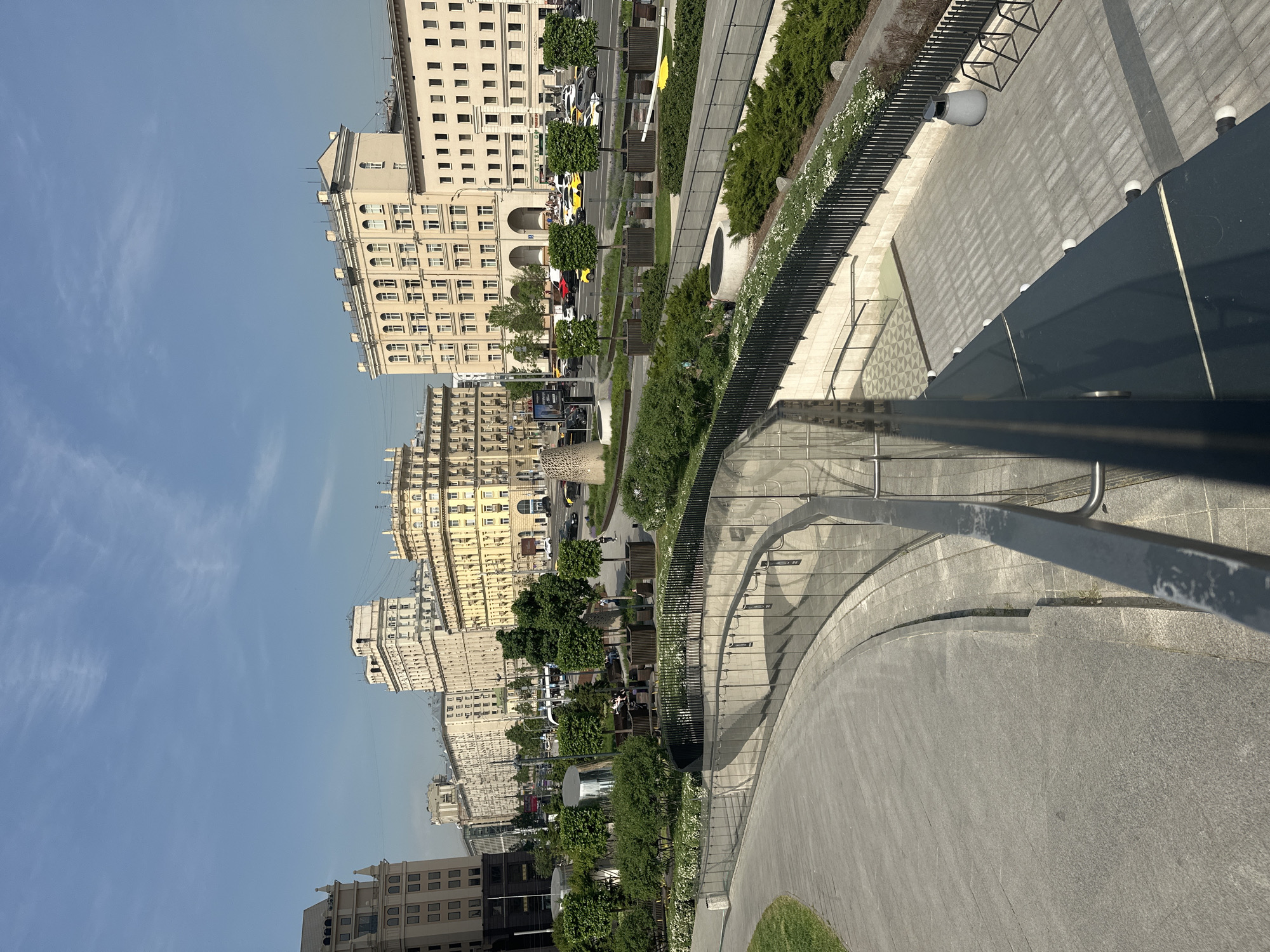 This image depicts an elevated view of Zaryadye Park in Moscow, Russia, including the surrounding cityscape. The park features tiered landscaping with walkways and benches, separated by rows of trees and shrubs. Pedestrians are seen sitting on the benches. Beyond the park, a line of multi-story, light-colored buildings stretches into the distance. A road with vehicular traffic runs parallel to the buildings. The sky above is a clear blue. The vantage point is from a high-level structure within the park itself, with part of its architectural railing visible in the foreground.