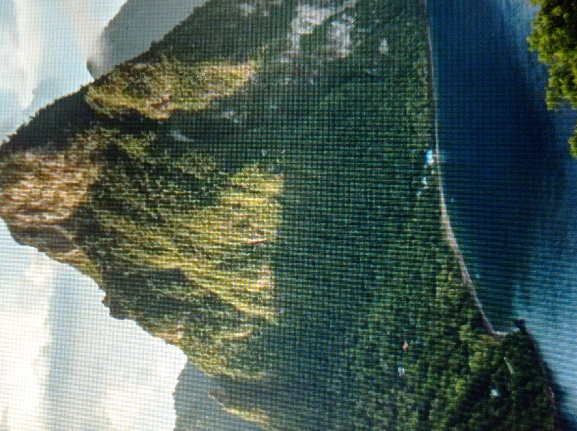 The image shows a lush, green mountain that slopes down to a dark blue body of water. The mountain is covered in dense vegetation and some rocky outcrops are visible. The water appears calm and reflects the light from the sky, which is partially cloudy. A thin strip of land with some trees and a few small buildings is visible along the edge of the water. The overall scene suggests a tropical or subtropical coastal environment. The location is not Mukachevo, Ukraine.