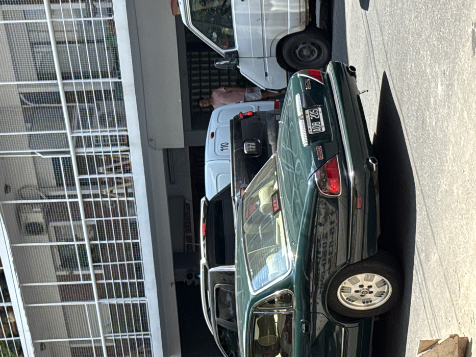 The image captures an outdoor street scene with several vehicles parked along what appears to be a curb next to a building. The perspective is slightly elevated, looking down the street. The lighting suggests bright daylight, casting strong shadows on the ground.
In the immediate foreground, positioned towards the right side of the frame, is the rear end of a dark green sedan. The car features a classic design with prominent taillights and chrome trim around the license plate holder. A "Jaguar" badge is visible below the left taillight. The license plate clearly displays "AOB 205". The car has twin exhaust pipes extending from underneath the bumper on the right side. Its wheels are multi-spoke alloy wheels. Reflections of the surrounding environment, including a building and possibly other vehicles, are visible on its glossy surface.
Behind the dark green sedan, moving further into the scene, are several other parked vehicles. Directly behind the Jaguar is a dark-colored vehicle, possibly a black SUV or another sedan, partially obscured. Following that is a white delivery van or utility vehicle. The number "110" is prominently displayed in a black oval on the side of this white van. It appears to be a Fiat Ducato or a similar model.
Further down the line of parked vehicles, a person is visible standing next to the white van. This individual is wearing a light-colored (possibly pink or peach) short-sleeved shirt and light-colored shorts. They appear to be an adult male. Behind this person and the white van is another, older white utility vehicle or small truck, partially visible.
To the left side of the frame, towering over the parked cars, is a large building with a distinctive white, grid-like facade. This grid structure, possibly made of metal bars or a patterned concrete, extends vertically and horizontally, suggesting either a multi-story building with grated windows or a large industrial structure. Through the grid, some interior details of the building can be vaguely seen, including what might be windows or internal structures. The overall scene conveys a typical urban or suburban street environment.