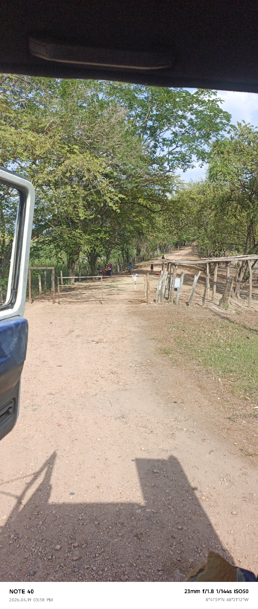 The image depicts a dirt road with trees on either side, taken from within a vehicle. The road has several barriers, one prominently in the foreground. In the distance, there are people present including children and a parked motorbike to the left. The environment is rural, with open sky visible through the canopy of trees. The location is indicated to be at 8°41'59"N, 68°21'12"W, which corresponds to the area around El Baúl, Venezuela. The shadow of a vehicle component and a hand wearing a glove are visible in the foreground.