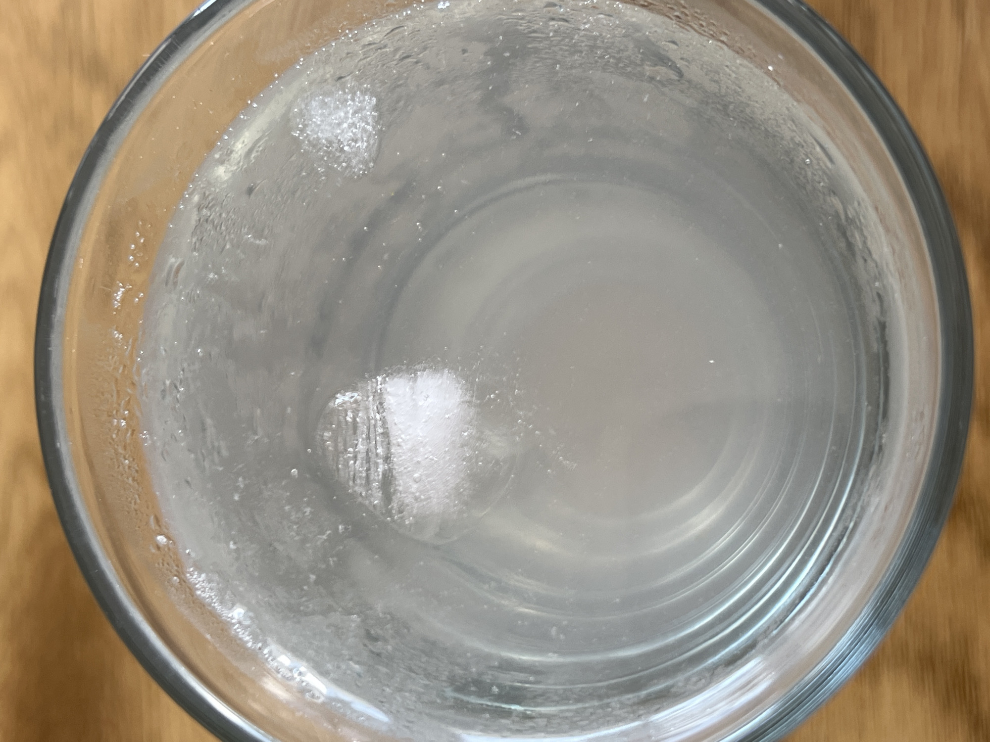 The image is a top-down view of a clear glass filled with a pale, cloudy liquid and ice cubes. The glass has condensation on the outside. The background is a wooden surface. There are no people or animals visible in the image. The image is not specific enough to determine a location in Mühldorf a. Inn, Germany.