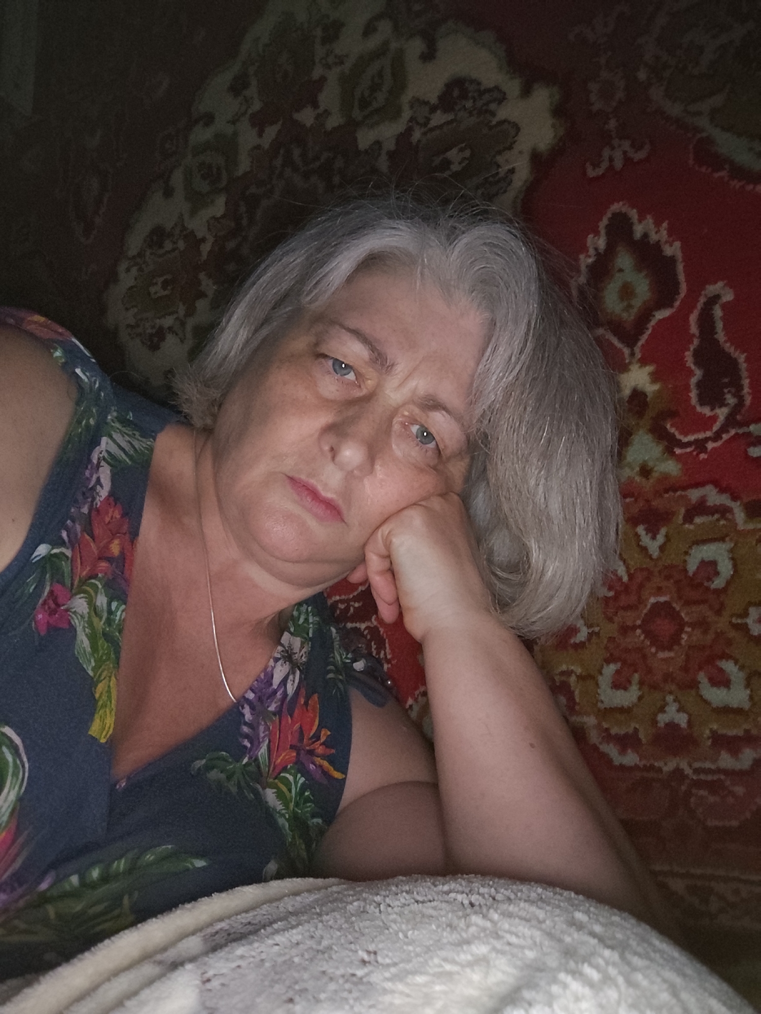 A middle-aged to elderly woman with gray, wavy hair and light blue eyes is depicted in a reclining position, resting her head on her right hand. She wears a dark blue sleeveless top or dress adorned with a multi-colored floral pattern, featuring red, orange, purple, and green elements. A thin silver chain necklace is visible around her neck. Her expression is neutral, with her gaze directed slightly downwards. The background consists primarily of a richly patterned rug or tapestry in shades of red, dark brown, and olive green, featuring ornate, traditional motifs. A portion of a white, textured blanket or towel is visible in the lower foreground, partially covering her lower body. The lighting is moderate, casting some shadows. The precise location within Krasnodar, Russia, cannot be determined from the visual information provided in the image.
